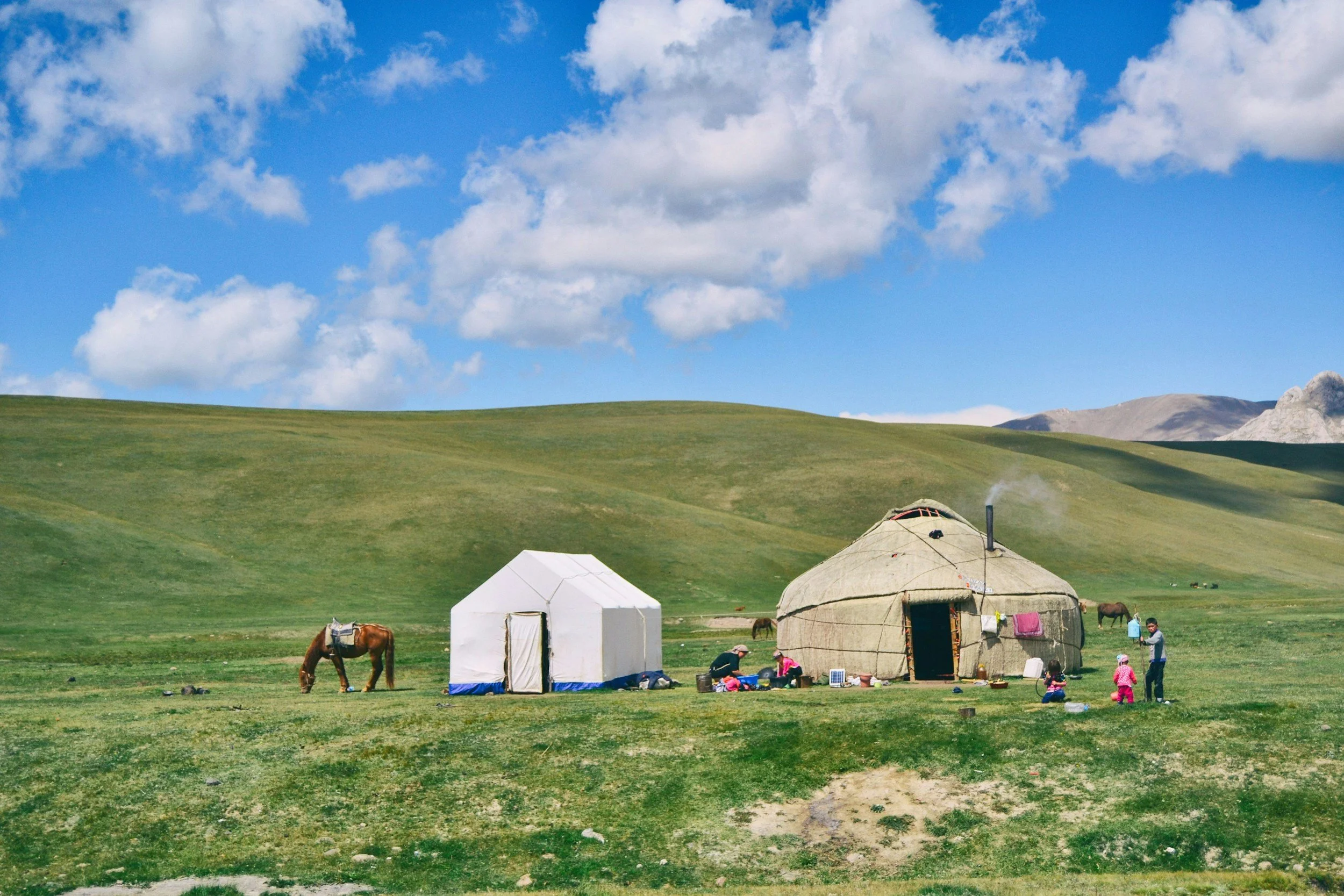 Scenic landscape with two tents and people outdoors, green rolling hills, a few horses grazing, and a partly cloudy blue sky.