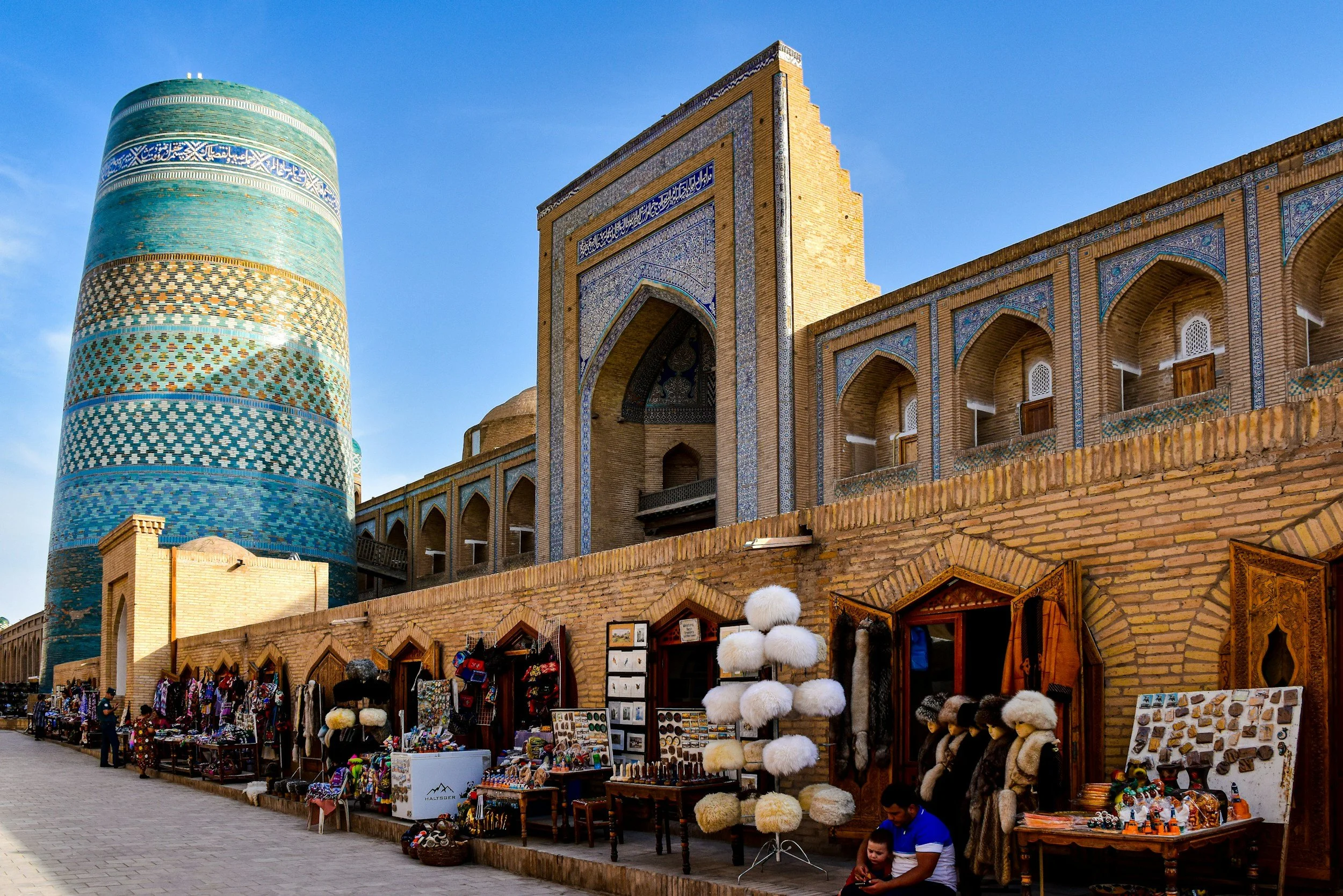 Market stalls selling souvenirs, hats, and jewelry in front of historical Islamic architecture with a tall, decorated minaret and ornate building facade, under a clear blue sky.