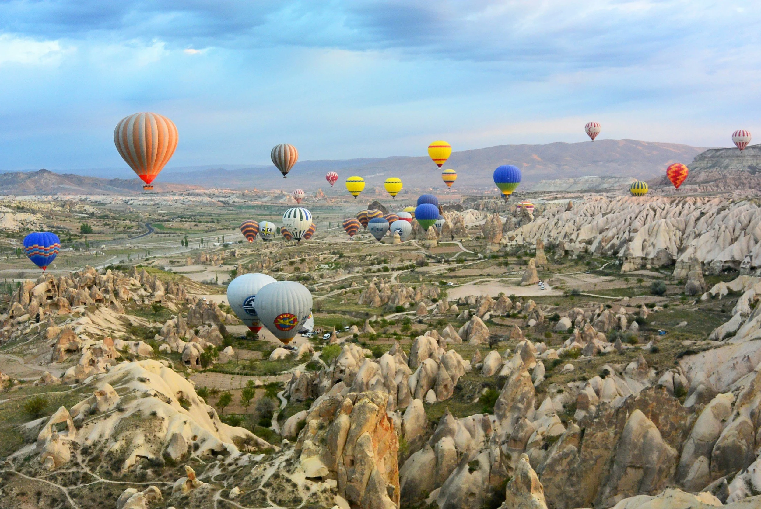 Multiple colorful hot air balloons floating over a rocky, mountainous landscape with valley roads and sparse vegetation.