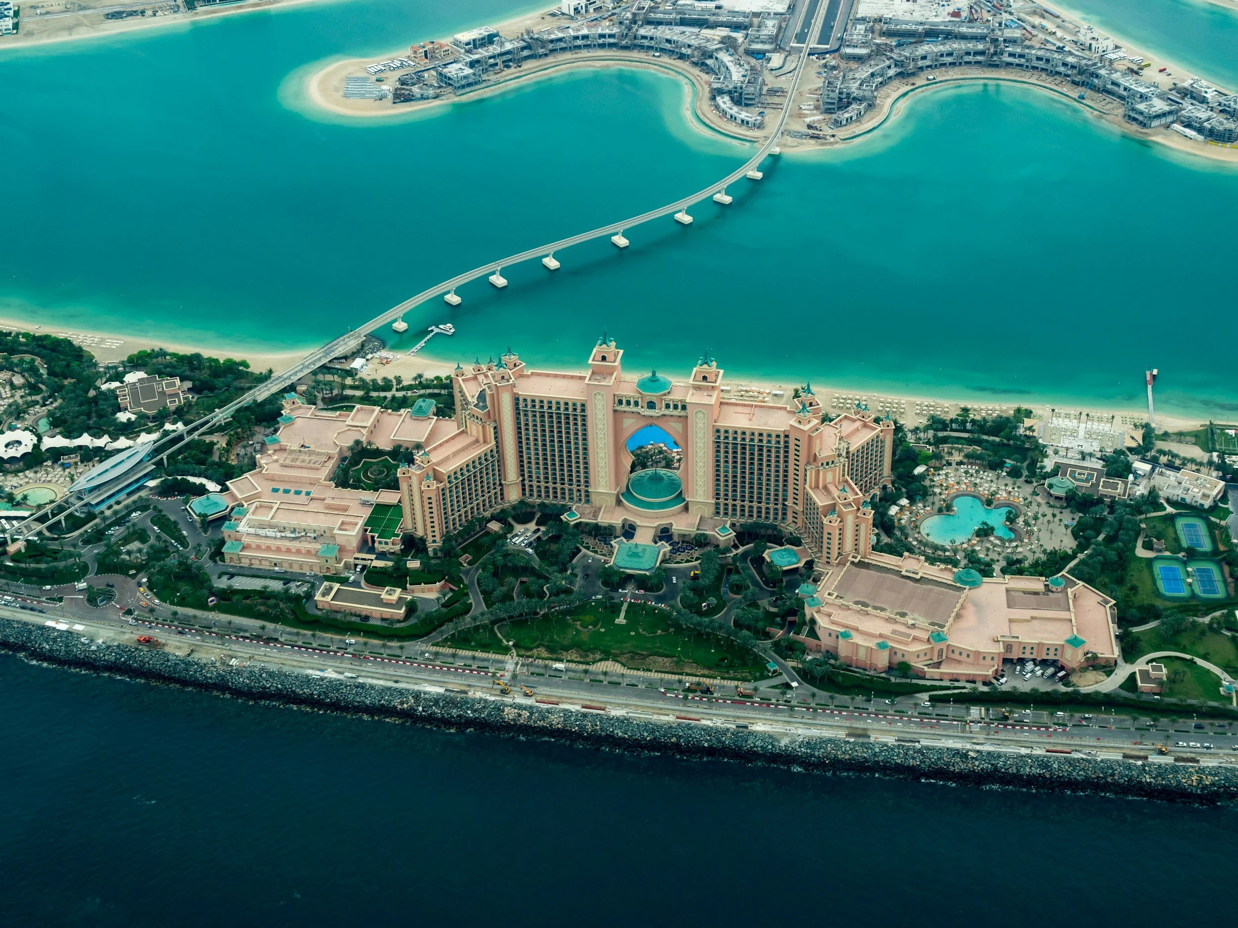 An aerial view of the Atlantis Paradise Island resort in the Bahamas, featuring a large pinkish hotel complex with towers, a bridge connecting to the island, multiple pools, tennis courts, and surrounded by beaches and turquoise waters.