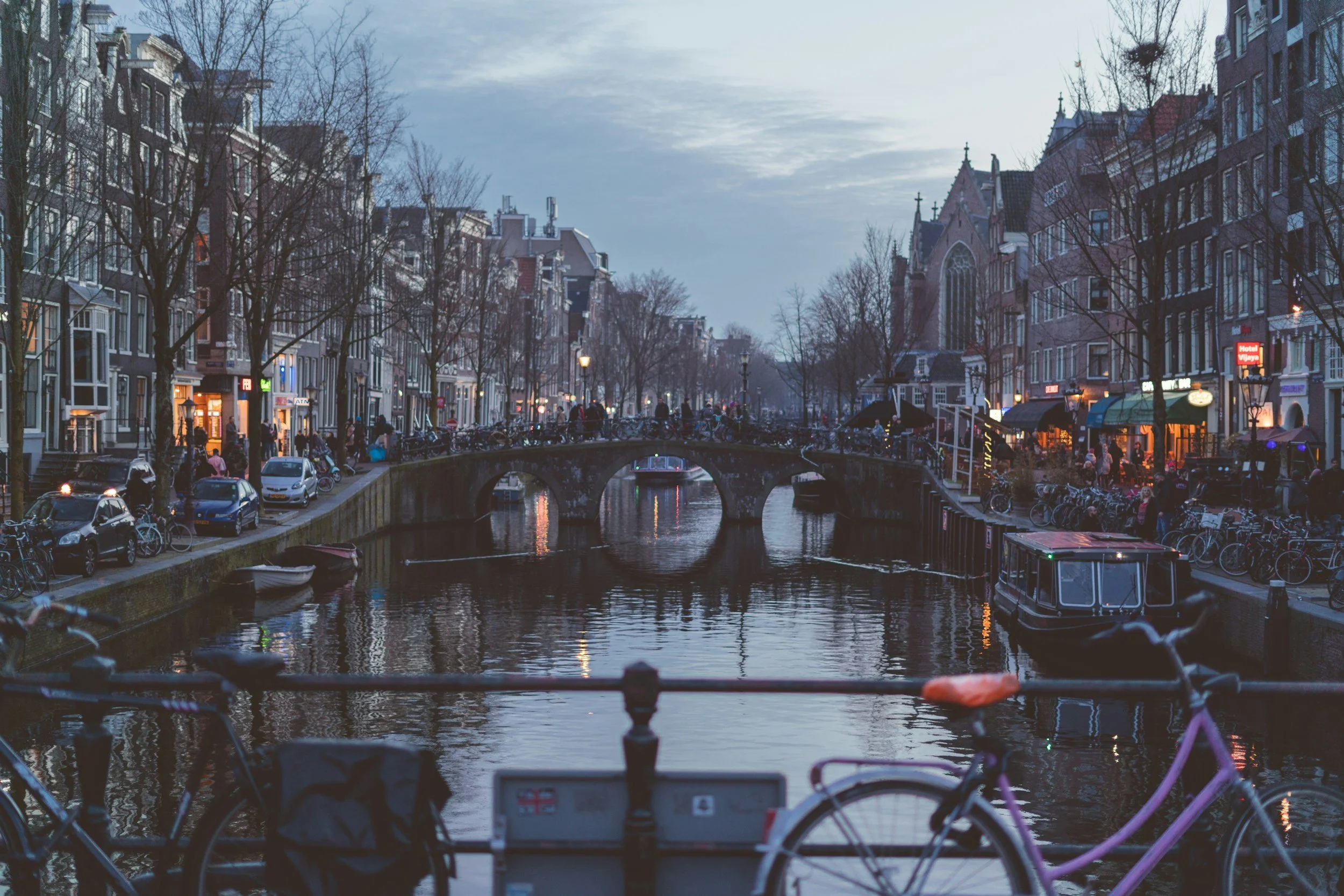 A canal scene in Amsterdam during twilight with boats on the water, bicycles on the foreground, and traditional Dutch buildings lining the canal with lights on.