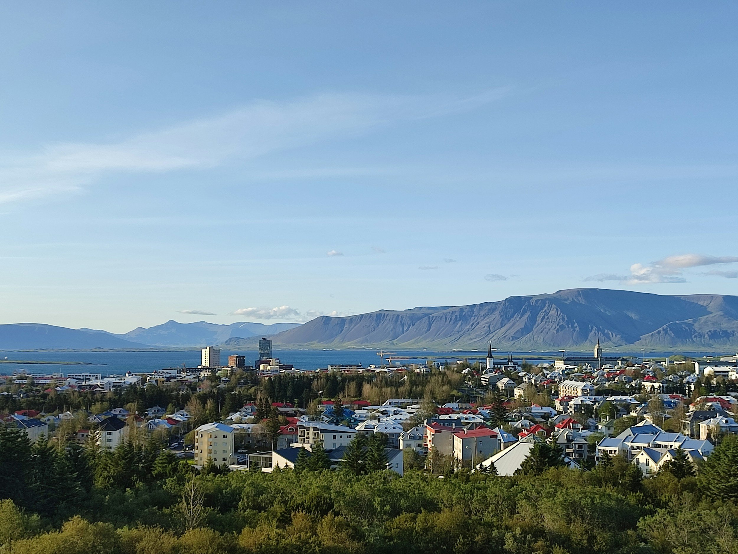 A scenic view of a coastal town with colorful houses, a few church steeples, and taller buildings across a body of water, with mountains in the background under a clear blue sky.