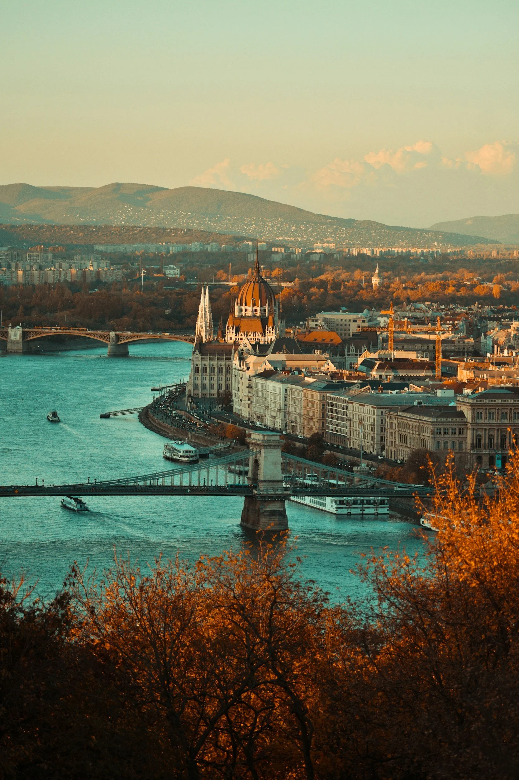 Aerial view of Budapest, Hungary, featuring the Chain Bridge, the Danube River, Parliament building, and Buda Hills in the background during sunset.