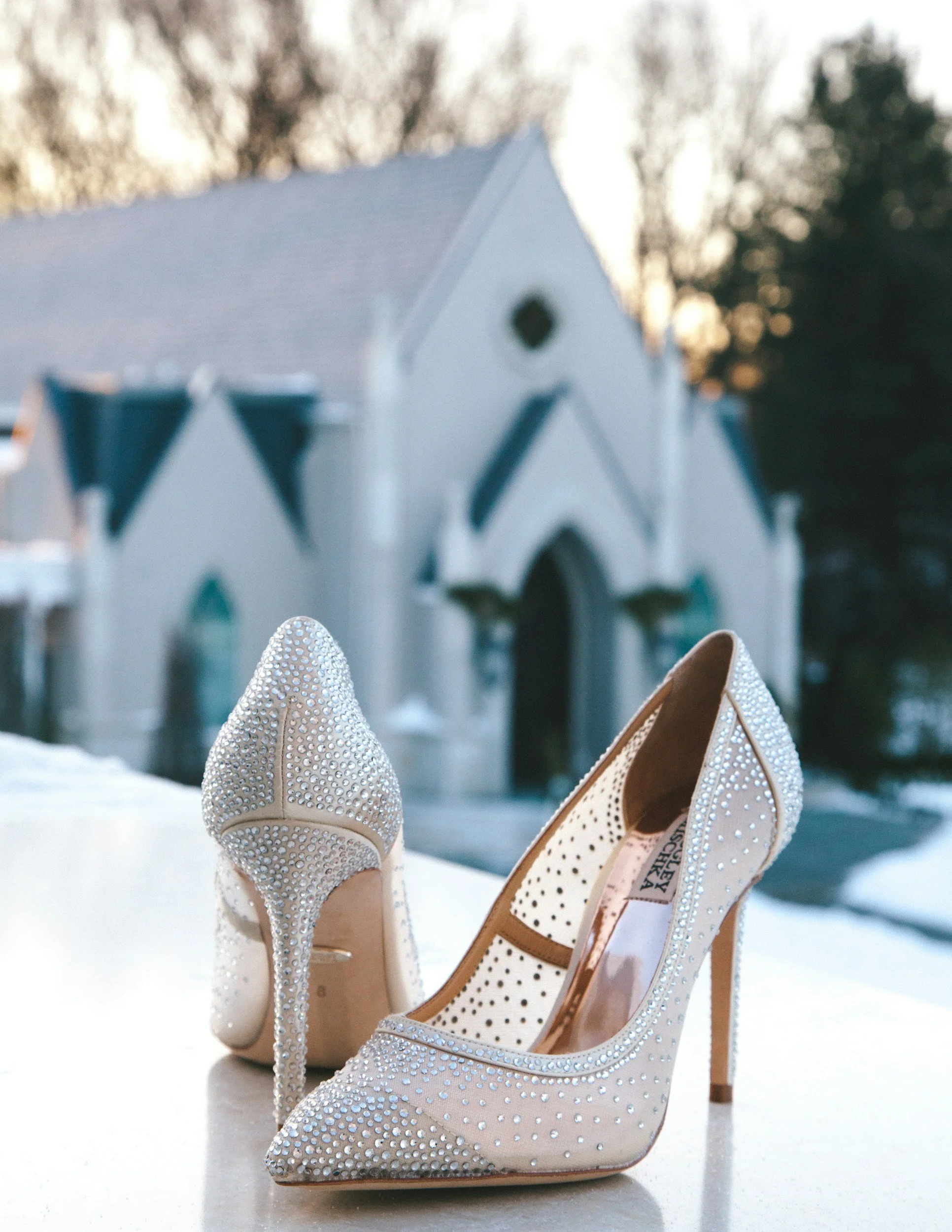 A pair of embellished high-heeled shoes with rhinestones, placed outdoors with a white church in the background.