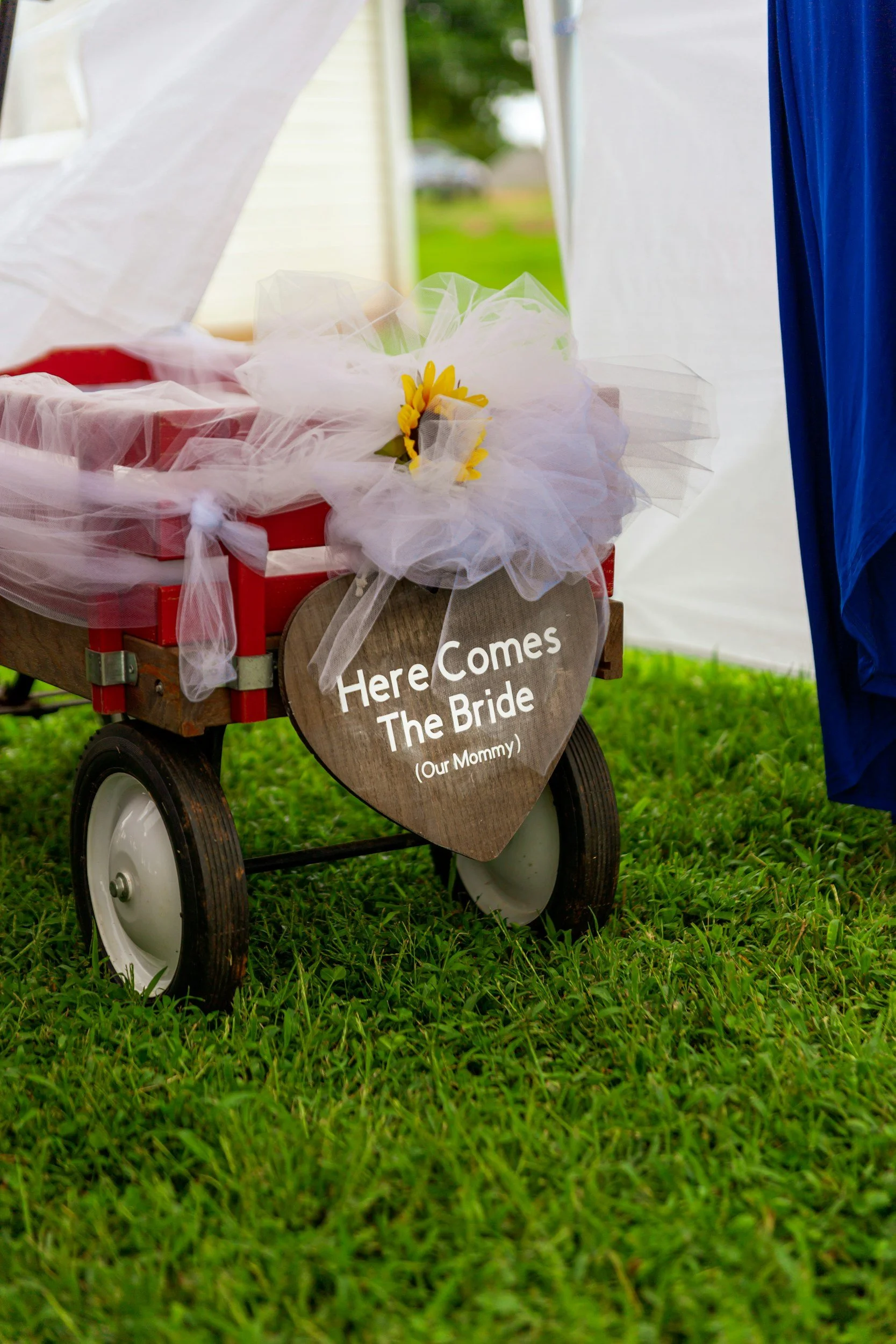 A red wagon decorated with tulle fabric, a sunflower, and a heart-shaped wooden sign with the text 'Here Comes The Bride (Our Mommy)'. The wagon is on a grassy outdoor setting, with a white tent and green trees in the background.