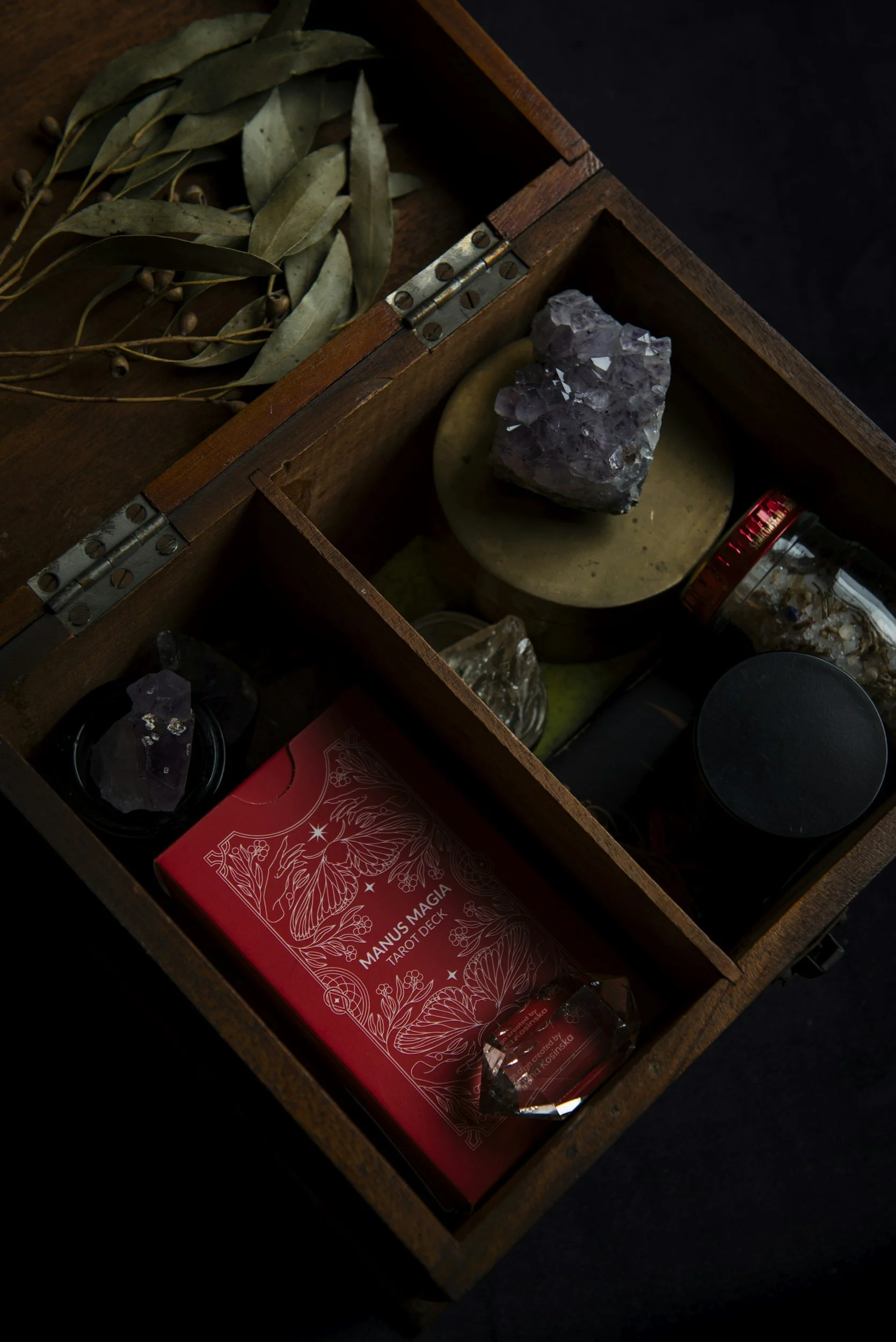 A wooden box containing dried leaves, crystals, a tarot deck titled "Manus Magia," and various small objects and containers.