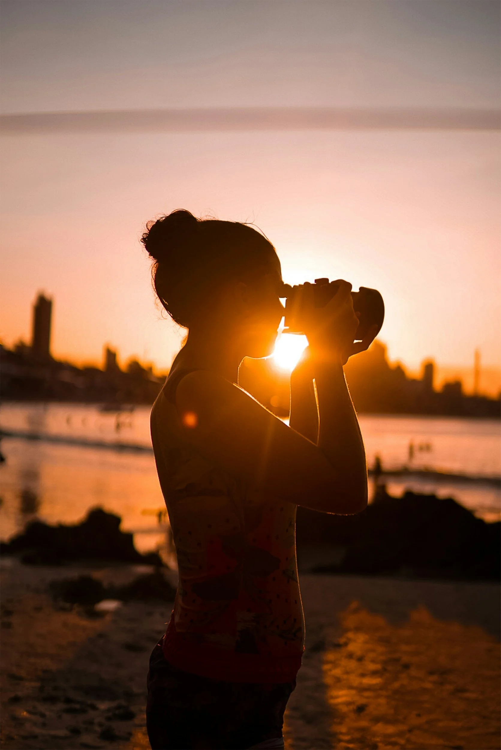 A person with their hair in a bun holding a camera takes a photo on the beach during sunset.