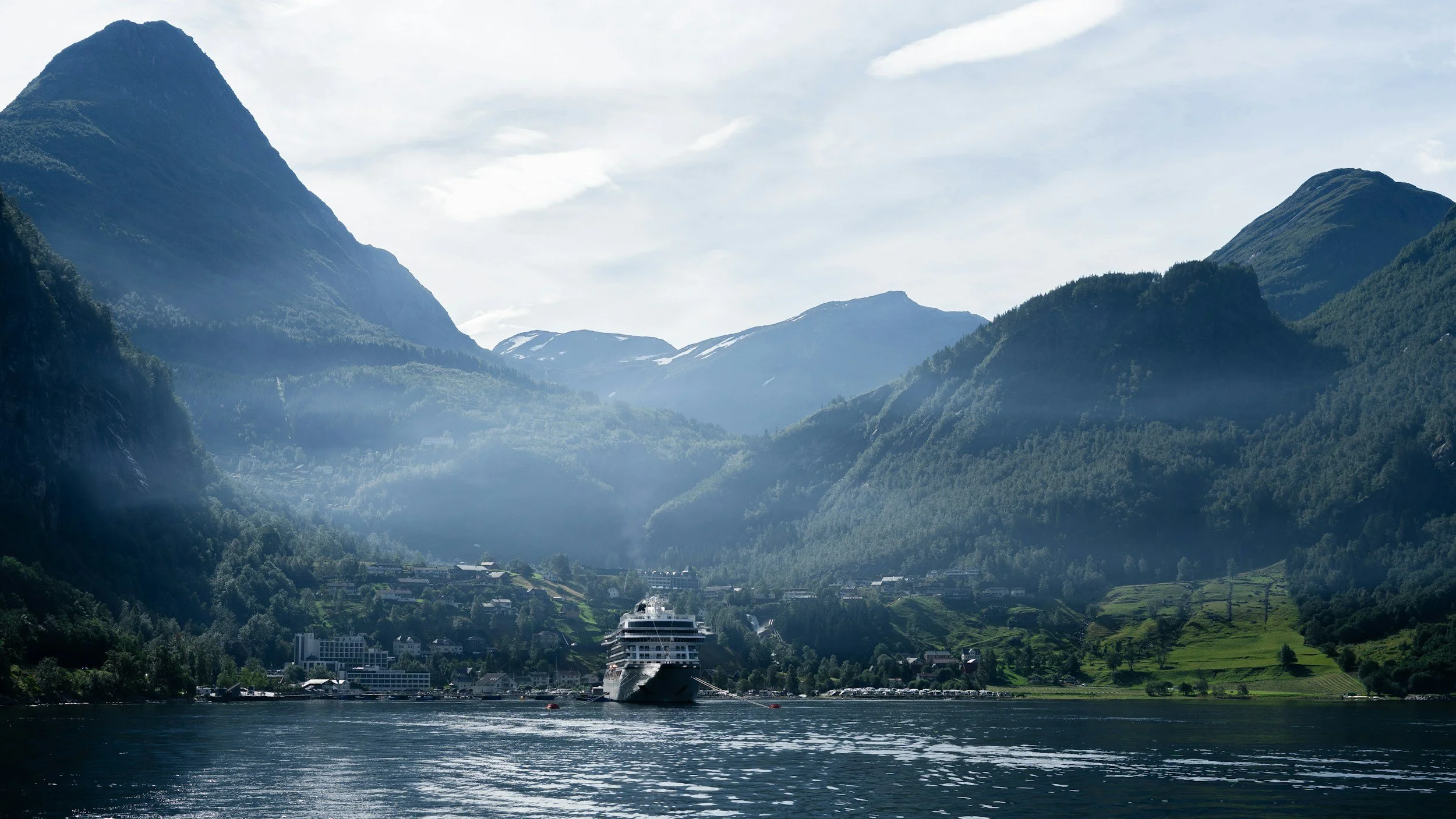 A cruise ship sailing on a body of water surrounded by towering green mountains with a small town at the foot of the mountains.