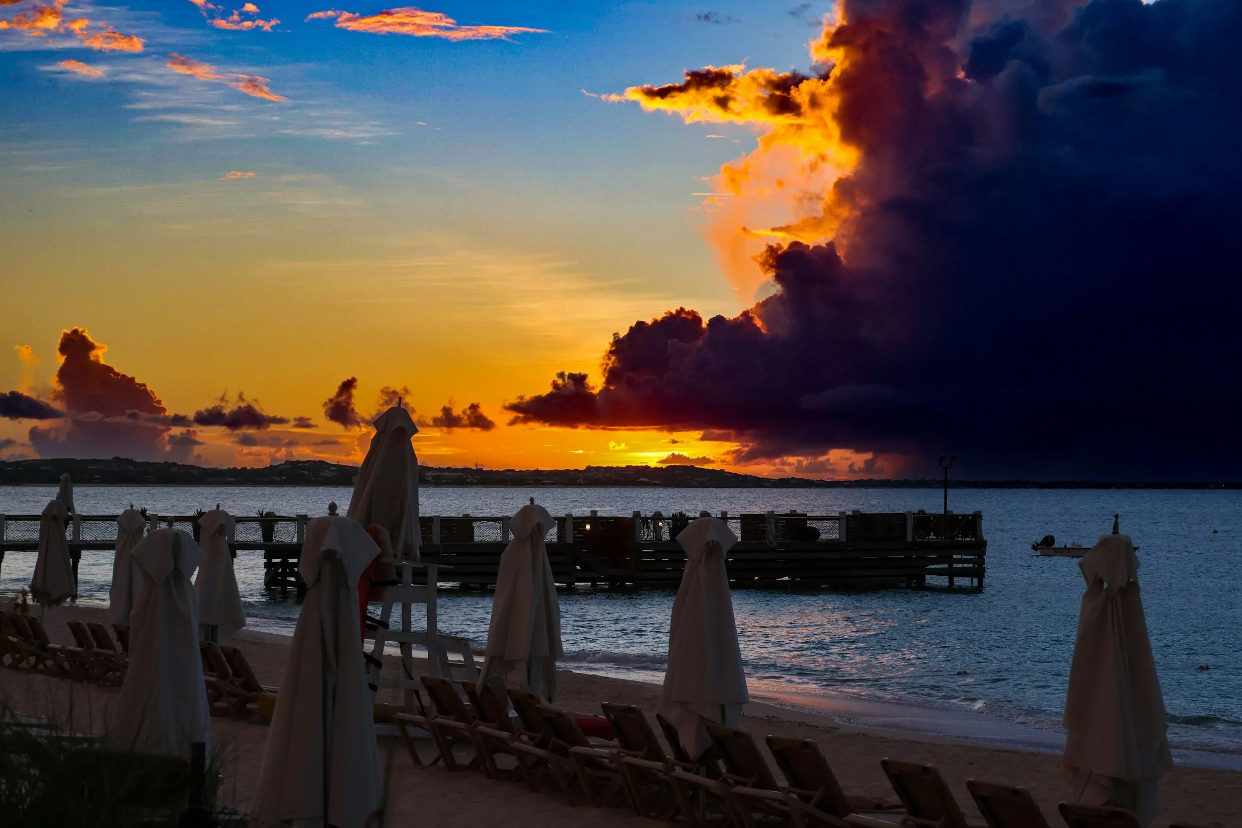 Sunset over the ocean with dark clouds and orange sky, beach chairs covered with white towels in the foreground, a dock extending into the water, and a small boat nearby.