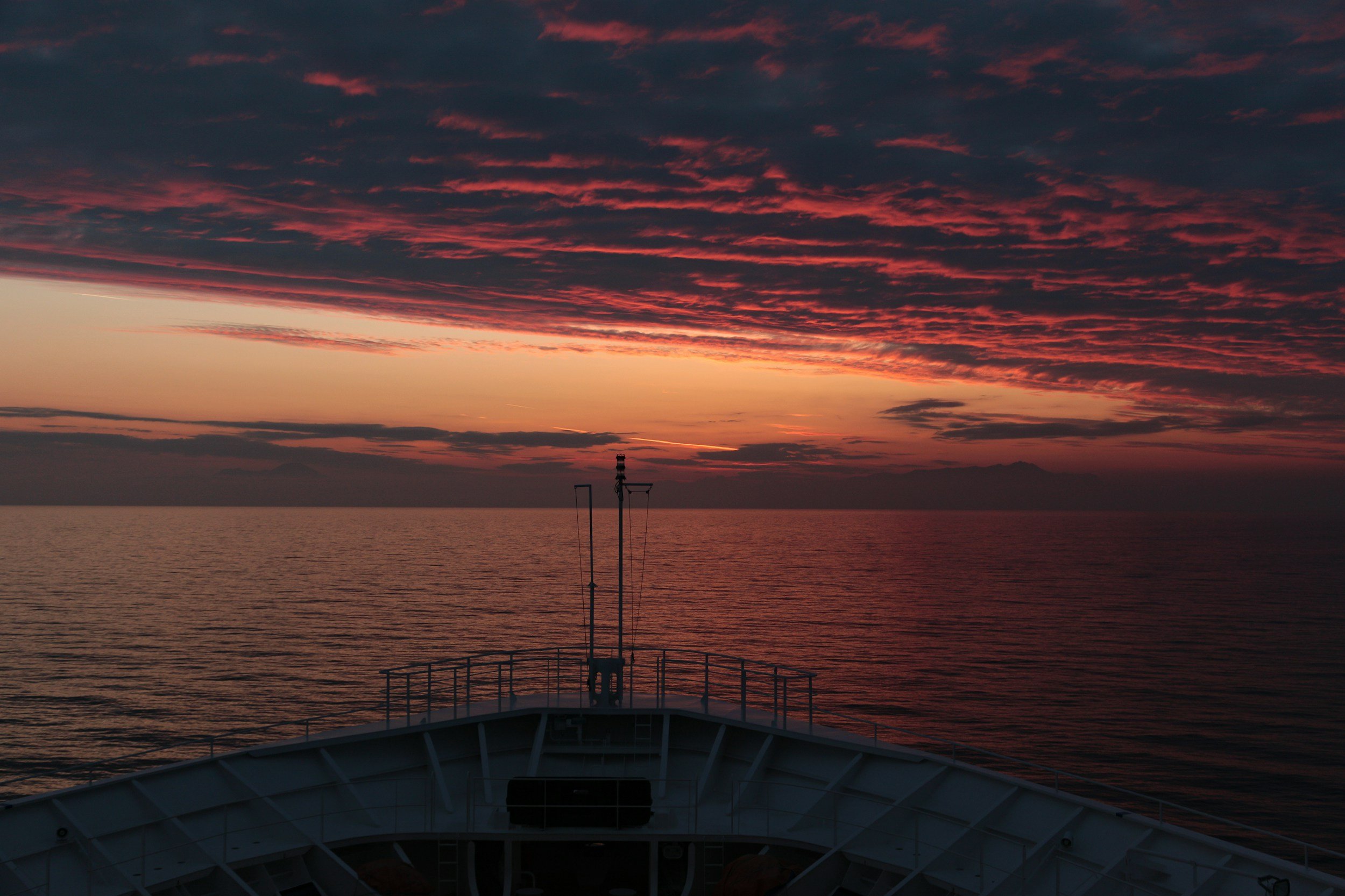 View from a boat's bow showing the ocean at sunset with a colorful sky of orange, pink, and purple clouds.