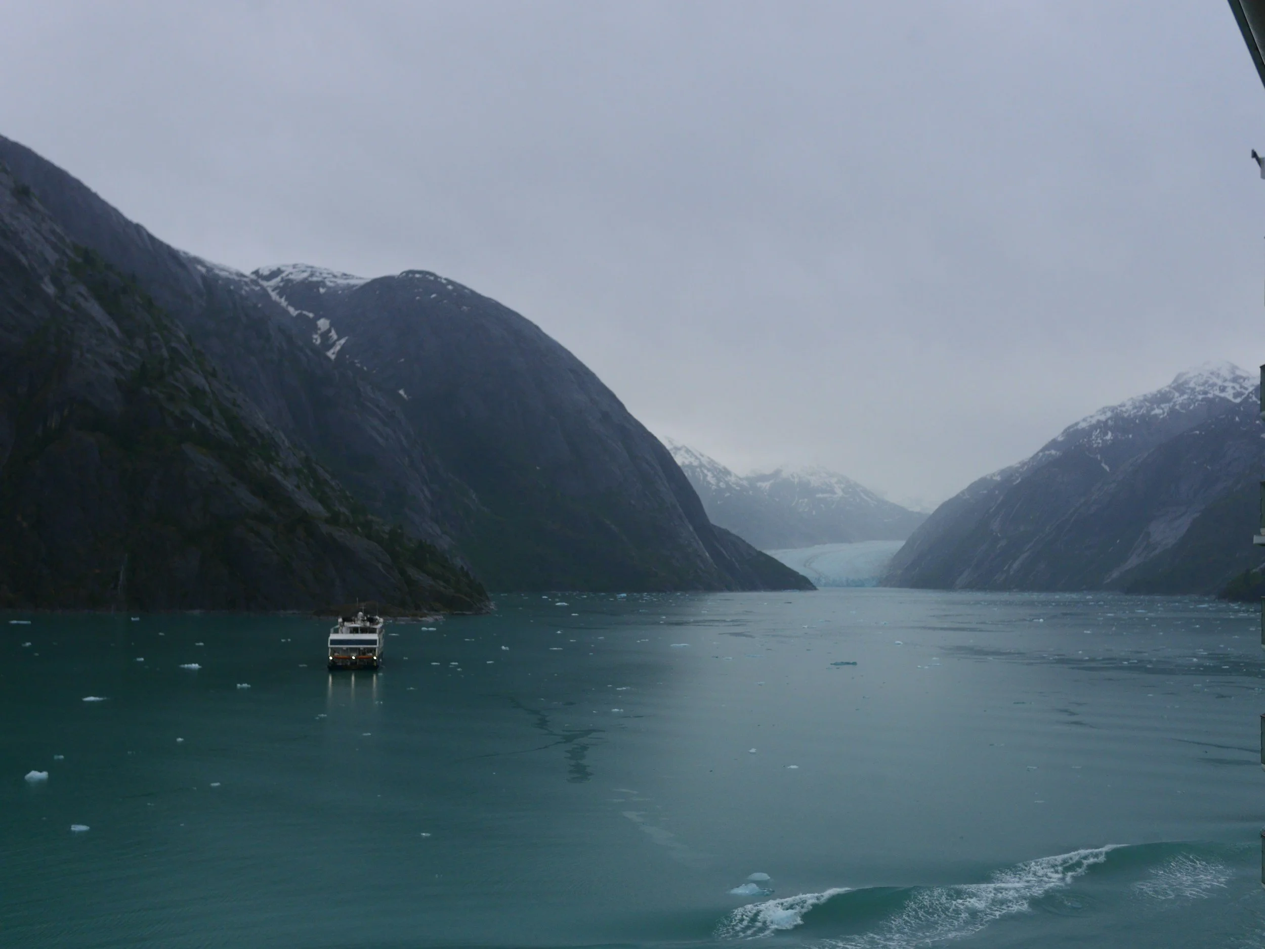 A boat sailing through a fjord surrounded by snow-capped mountains on a cloudy day.