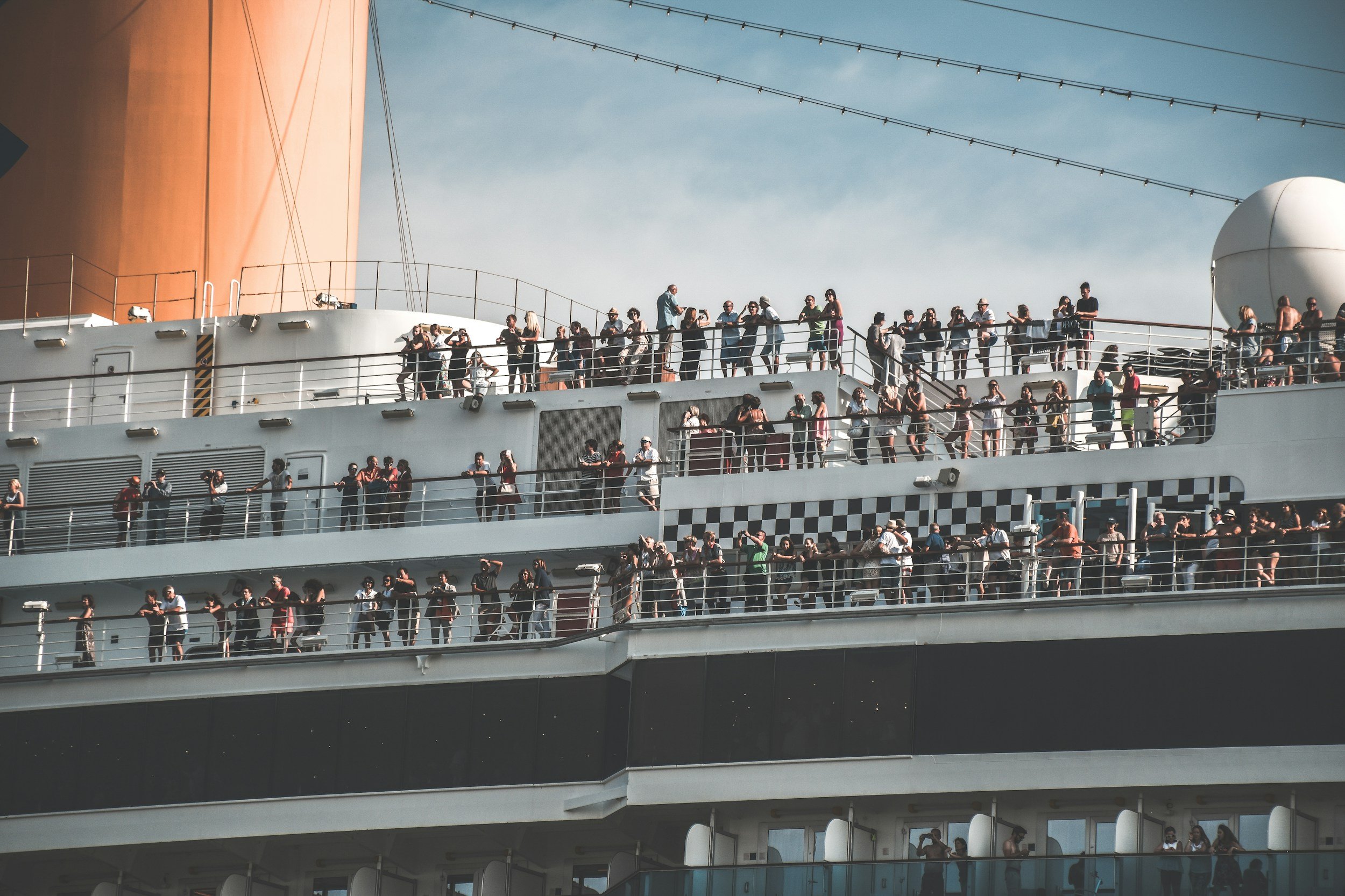 Crowds of people on the outdoor deck of a cruise ship, some taking photos, with blue sky and a large orange and white structure in the background.