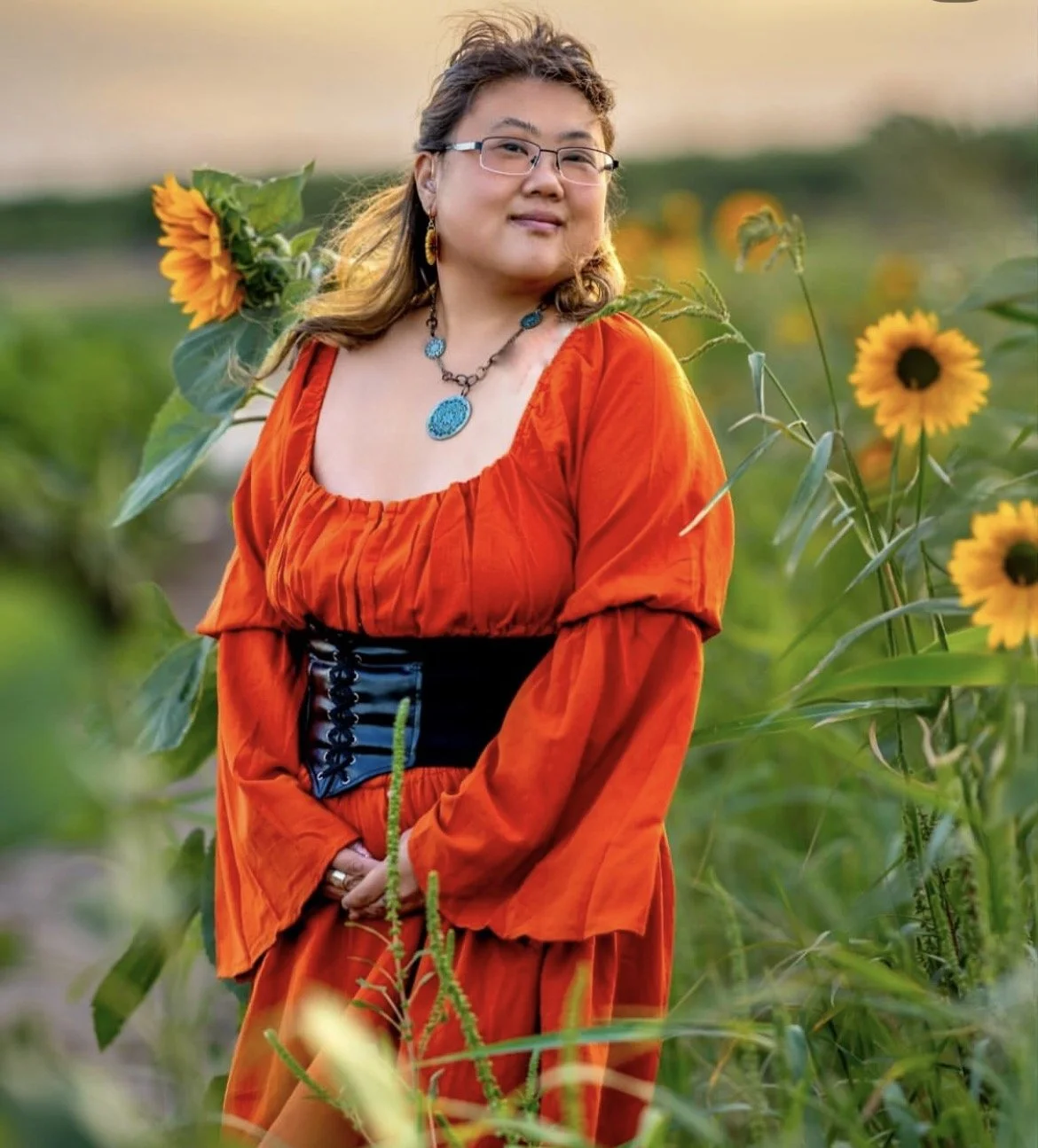 A woman wearing an orange dress with a black corset standing in a sunflower field during sunset, with mountains in the background.