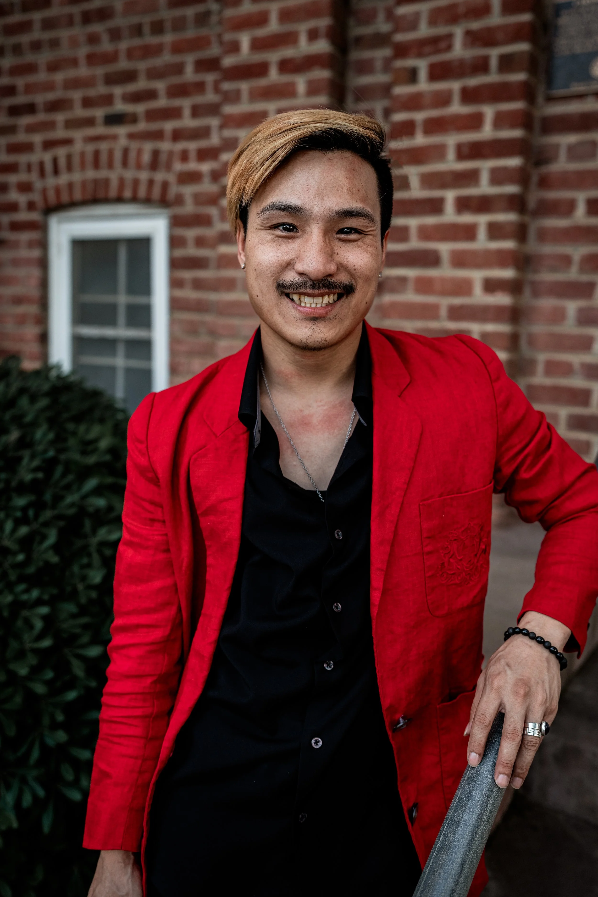 A smiling man with light brown and black hair, (possibly Asian descent) wearing a red blazer over a black shirt, standing outdoors near a brick building with a window and green shrubbery.