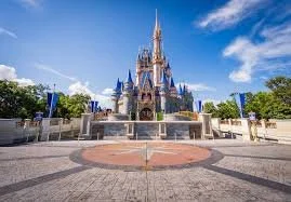 Disney's Cinderella Castle at Walt Disney World in Orlando, Florida, under a blue sky with clouds, surrounded by trees and a paved area.