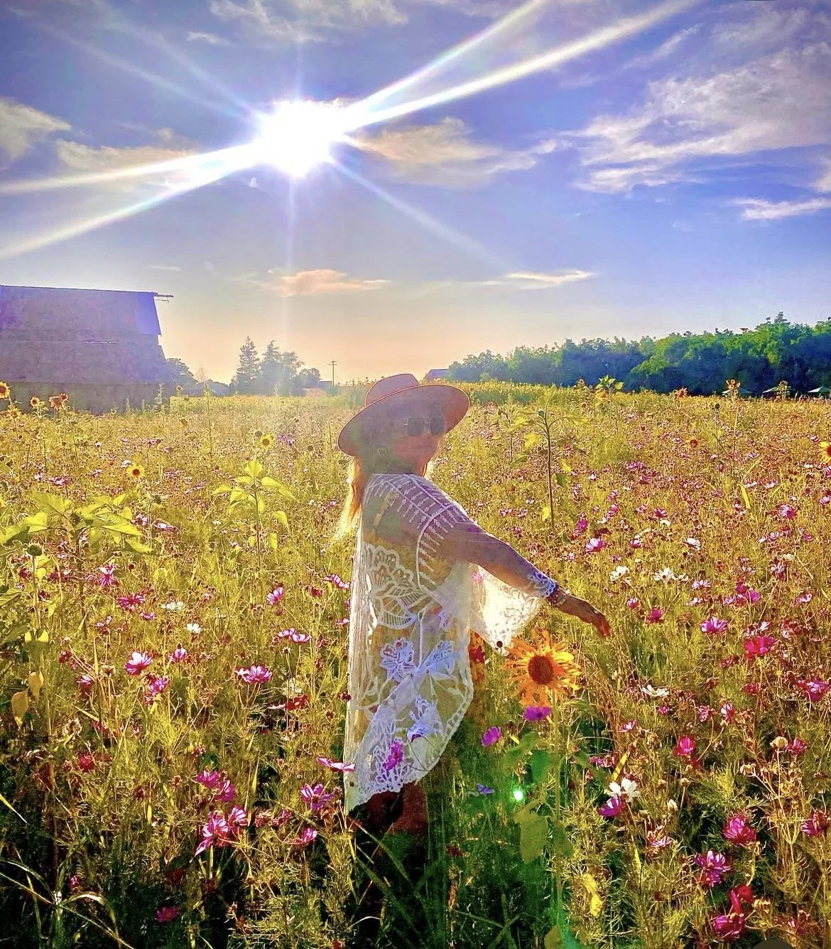A woman in a white lace dress and a wide-brimmed hat standing in a colorful flower field under a bright sun with a rainbow-like halo.