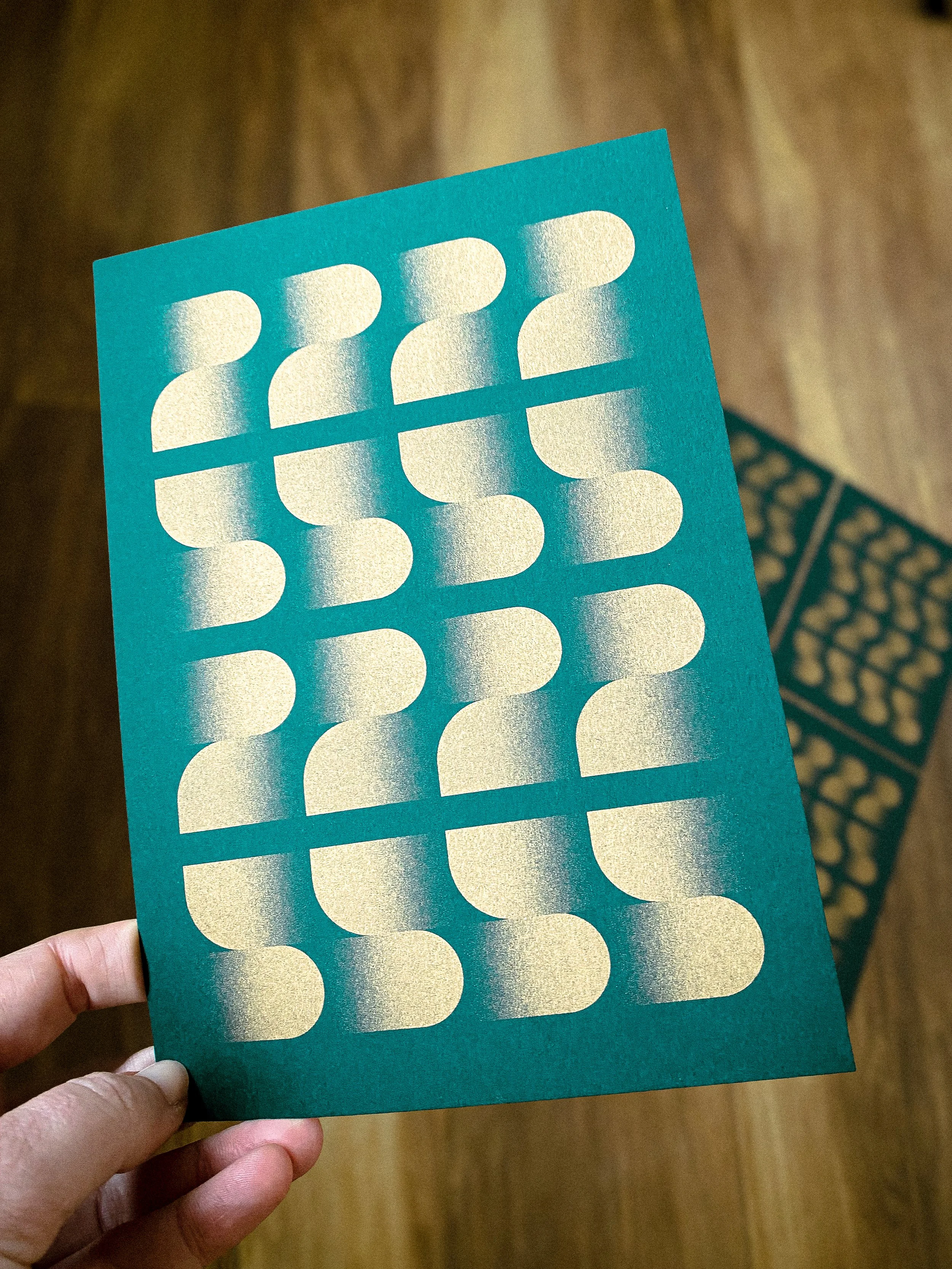 Une main tient une carte de vœux avec des motifs dorés en forme de demi-cercle, disposés en rangées.