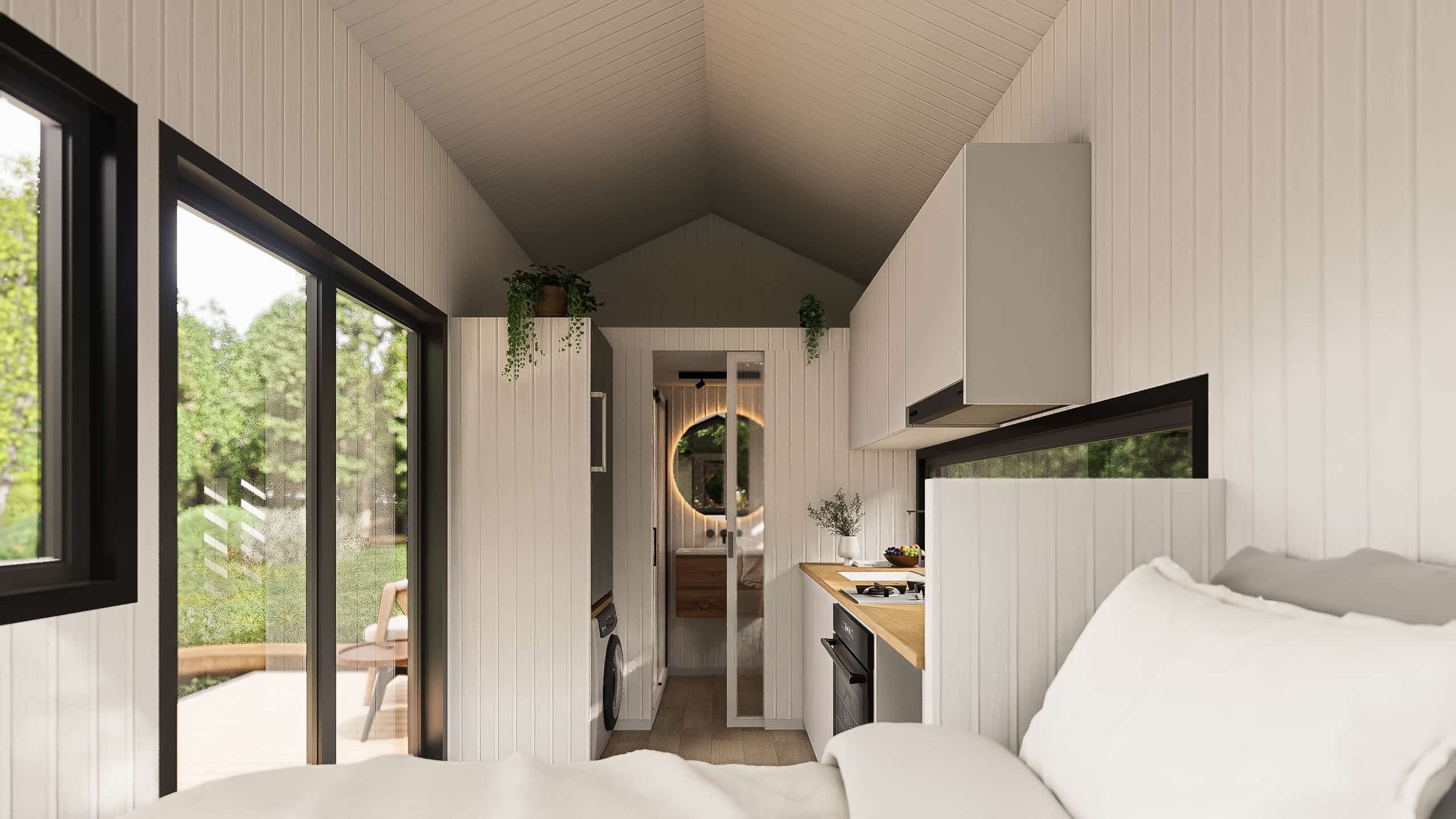 Interior of a small, modern tiny home with white paneled walls, a bed in the foreground, a kitchen with a wooden countertop, and a sliding door leading to an outdoor patio with a chair and green trees outside.