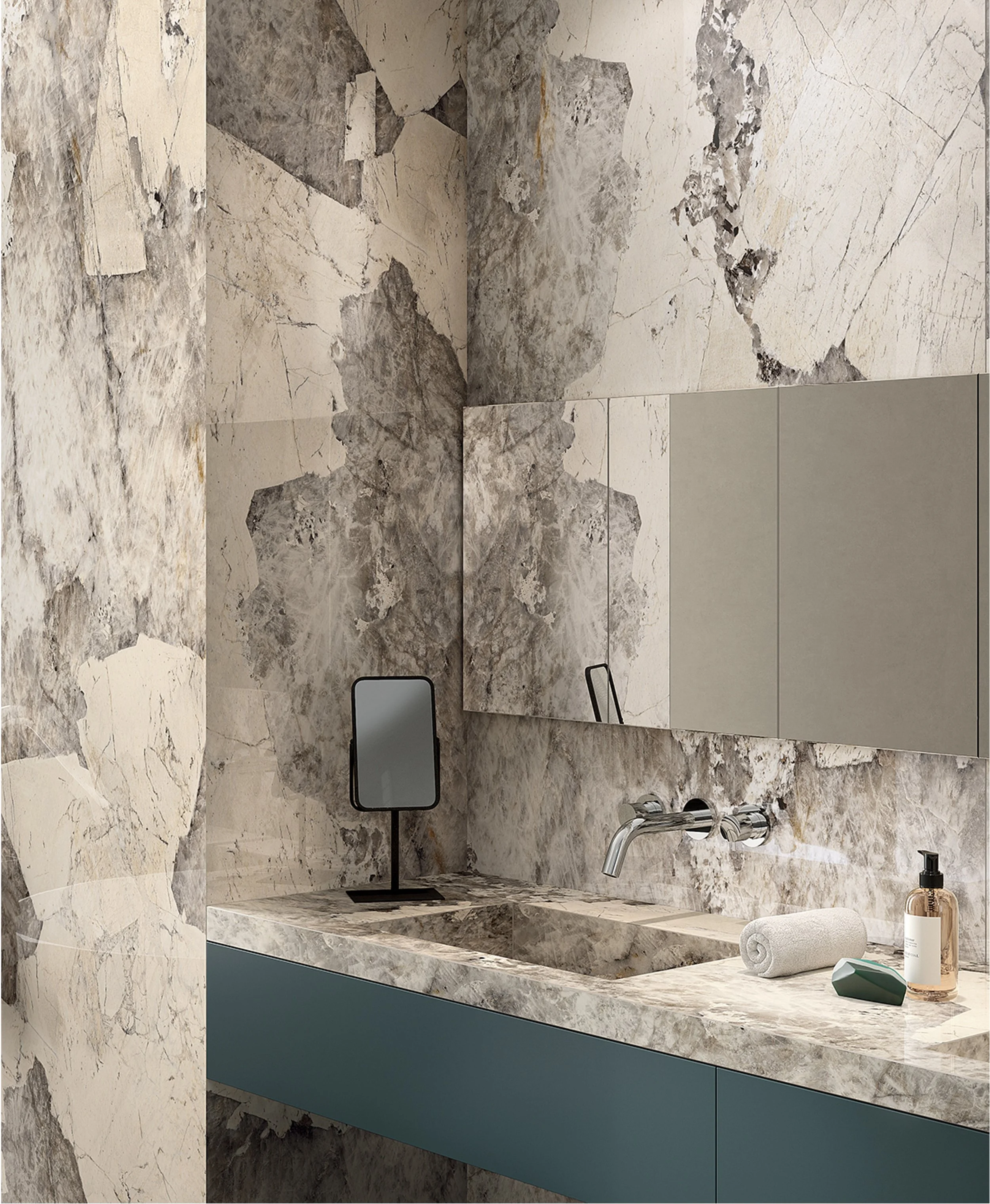 Modern bathroom with marble walls and countertop, mirror, small stand-up mirror, rolled towel, soap dispenser, and small decorative objects.