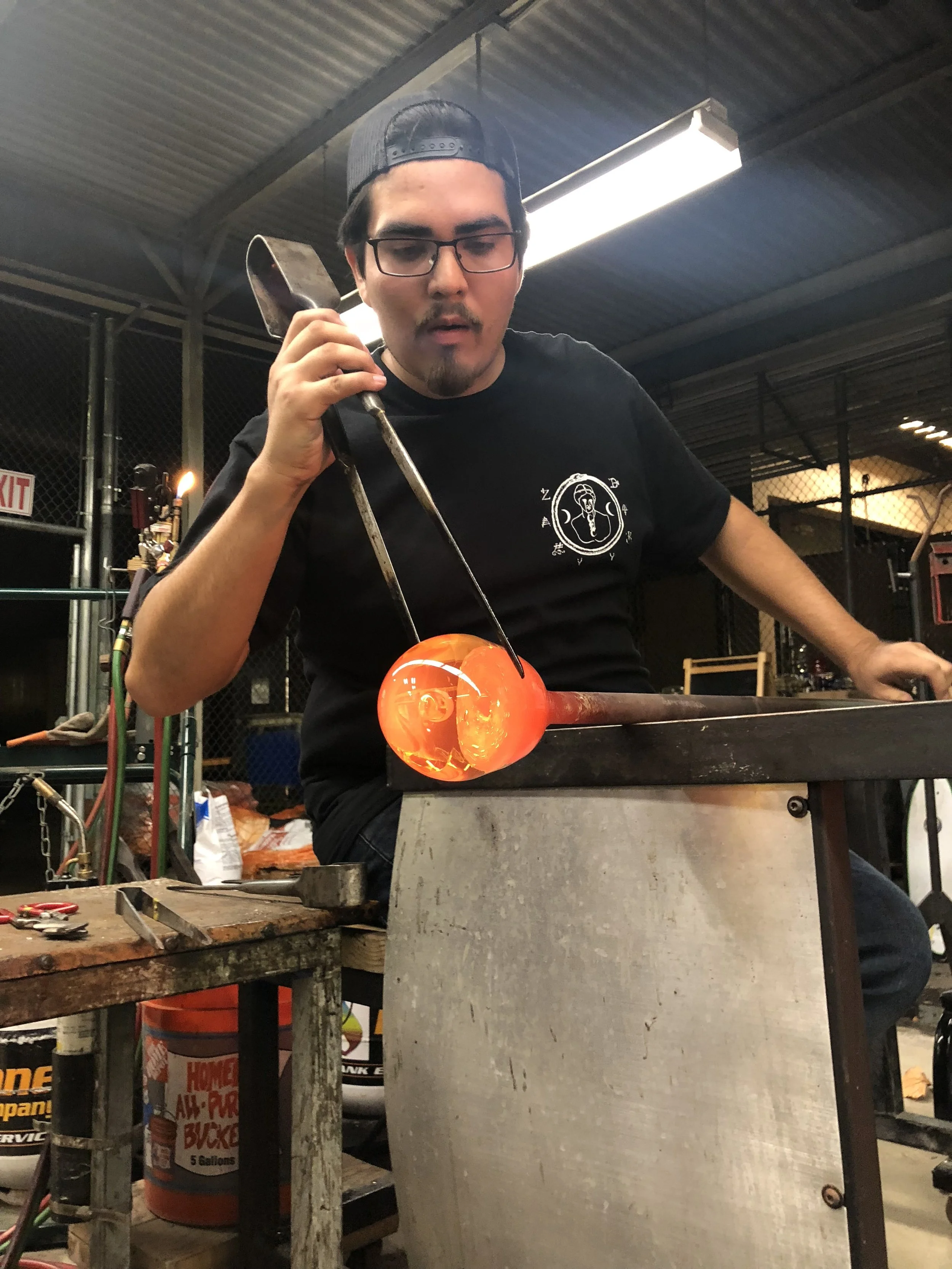 A person wearing glasses and a black cap is working on a glassblowing project at a workbench, shaping a glowing orange molten glass piece with tools in an industrial workshop.