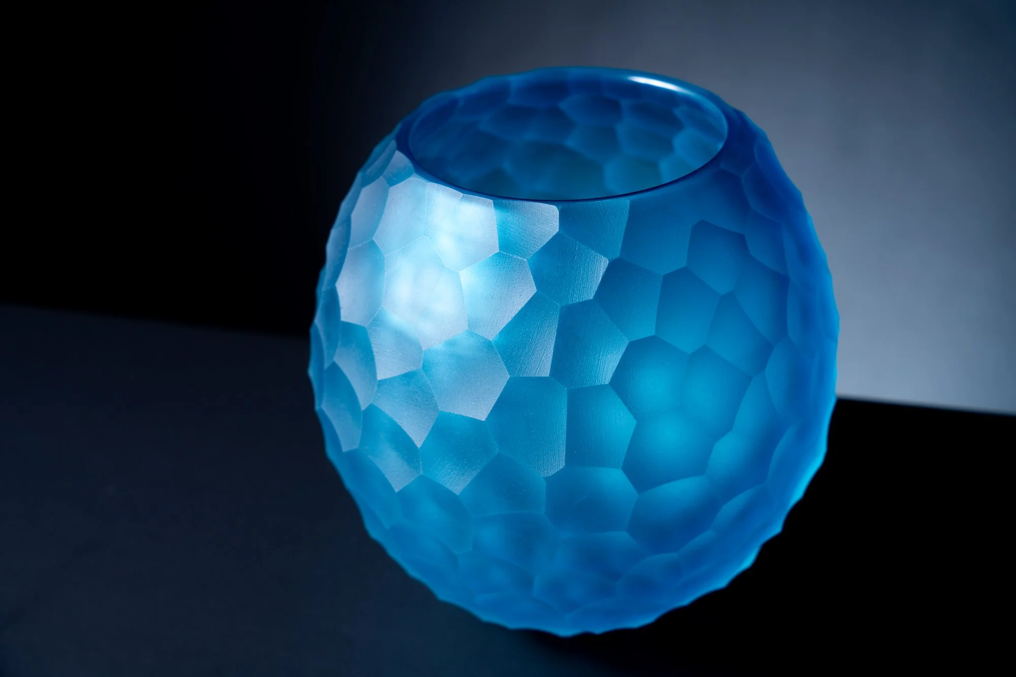 A close-up of a blue, textured, geometric light fixture with a honeycomb pattern, reflecting on a glossy surface beneath it.