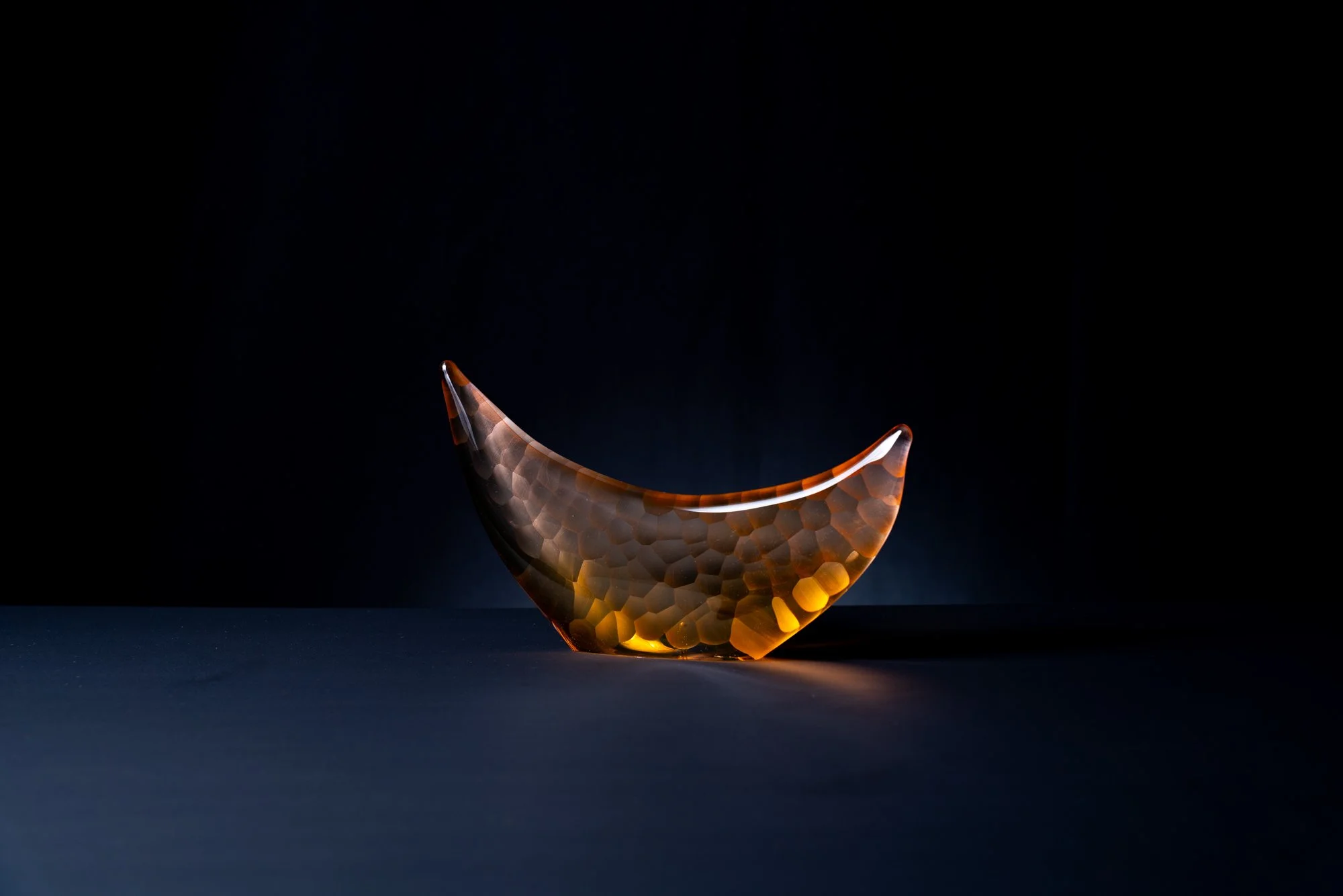 A glass teardrop-shaped sculpture with a honeycomb pattern glowing with amber light, set against a dark background.