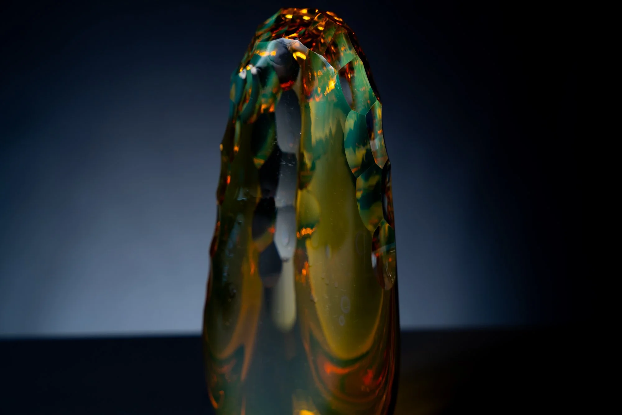 Close-up of a multicolored, translucent, faceted glass sculpture with a reflective surface, against a dark background.