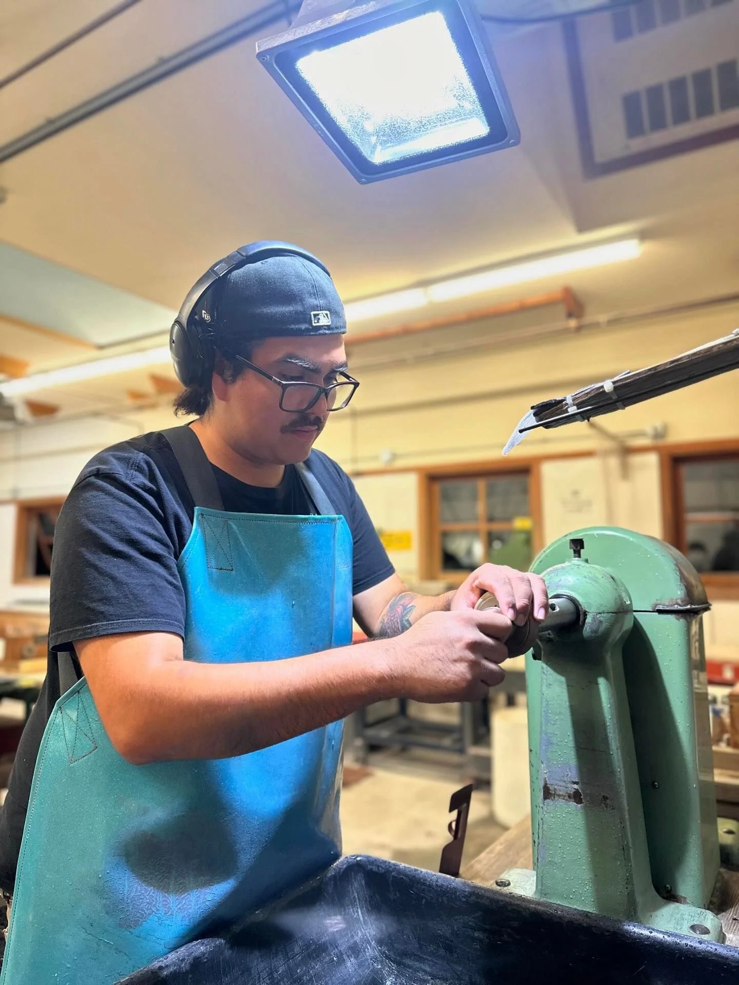 A man wearing safety glasses, a dark cap, and a blue apron working with a green metal machine in a woodworking shop.