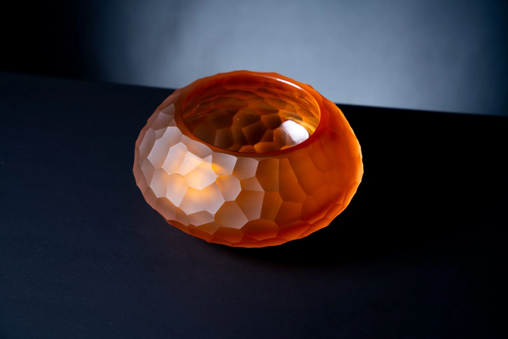 An orange, faceted, spherical lamp with a honeycomb-like pattern, placed on a reflective black surface with a plain dark background.