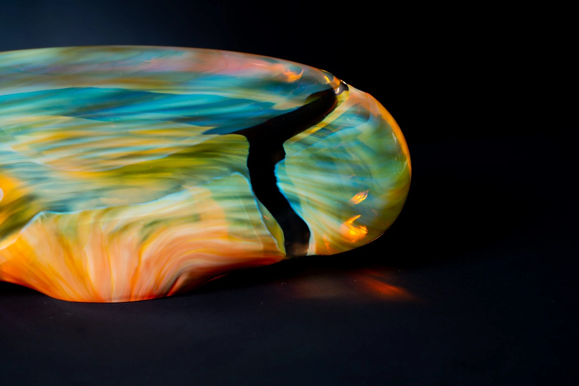 Close-up of a colorful, artistic glass object with swirling patterns in shades of blue, yellow, orange, and black, set against a black background.