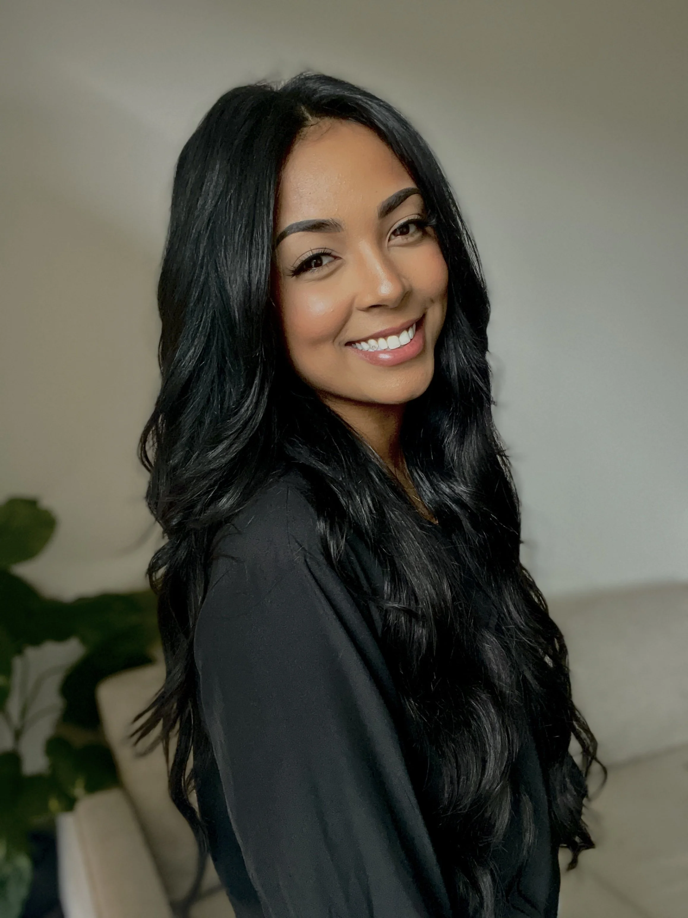 A smiling woman with long black hair, wearing a black top, standing indoors with green plants in the background.