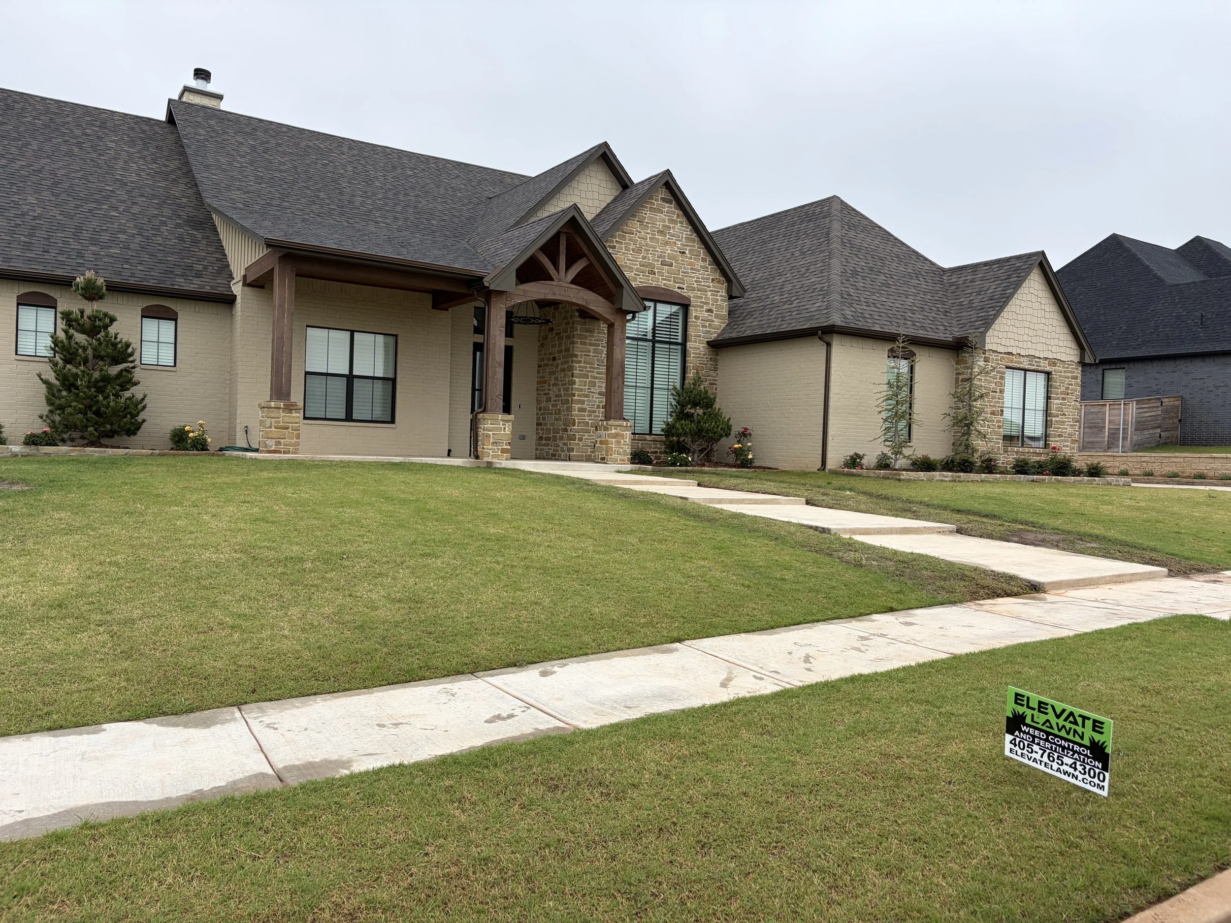 Large stone-and-brick suburban house with a covered entry, manicured lawn, sidewalk, and weed control sign