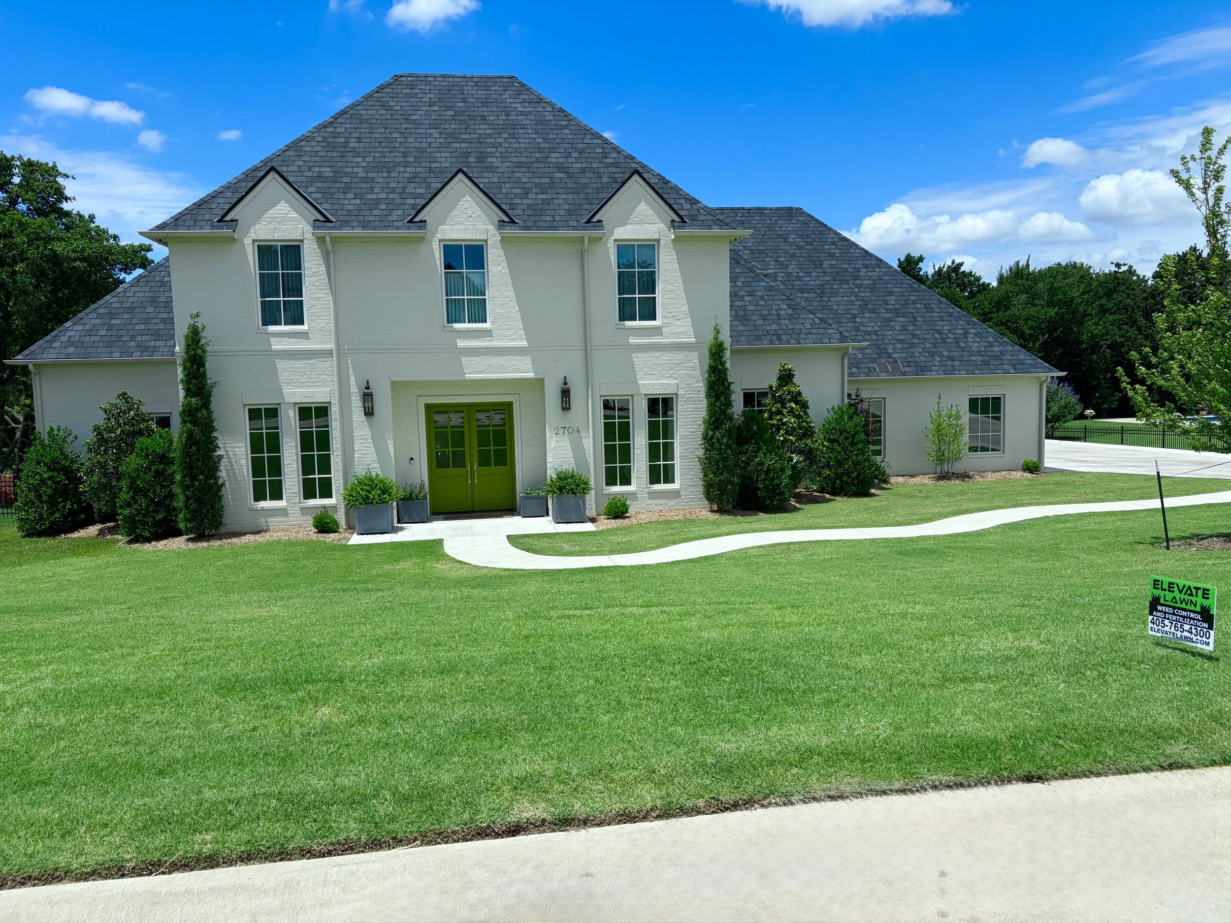 Image of a large white house with green door and tall windows, surrounded by green lawn and trees, under a blue sky with some clouds. There is a weed control sign in the yard.