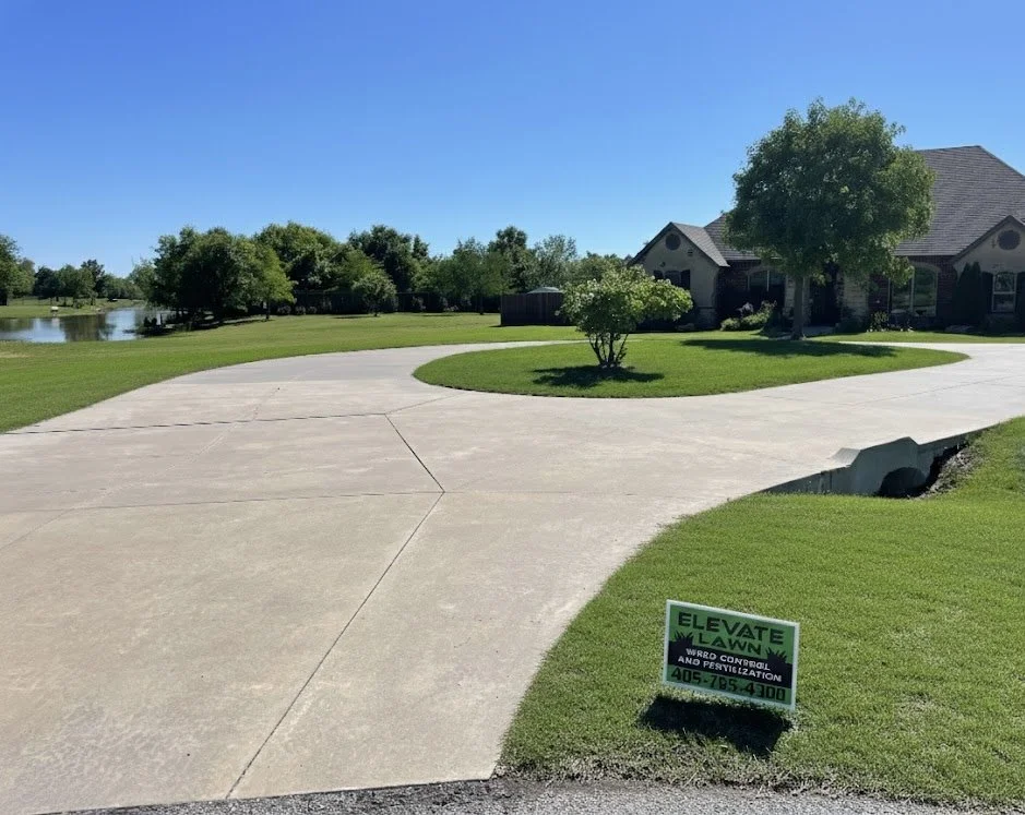 Concrete driveway curves in front of a house with a well-kept lawn, trees, and a pond in the background under a clear blue sky.
