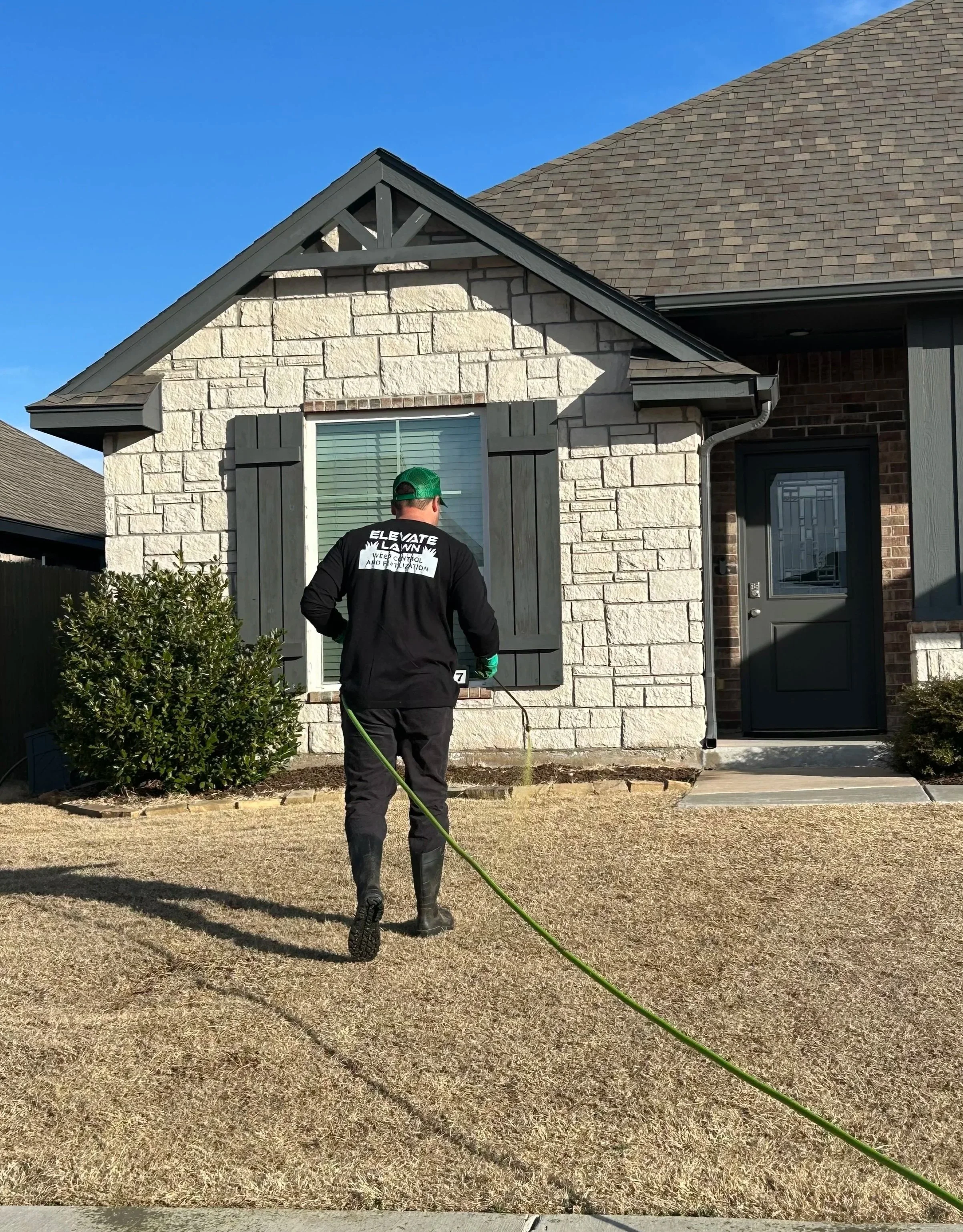 A person wearing a green cap, black jacket, and boots is spraying pesticide or water on the front yard of a house with a garden hose. The house has a gray door, brick and stone exterior, dark gray shutters, and a small shrub nearby.
