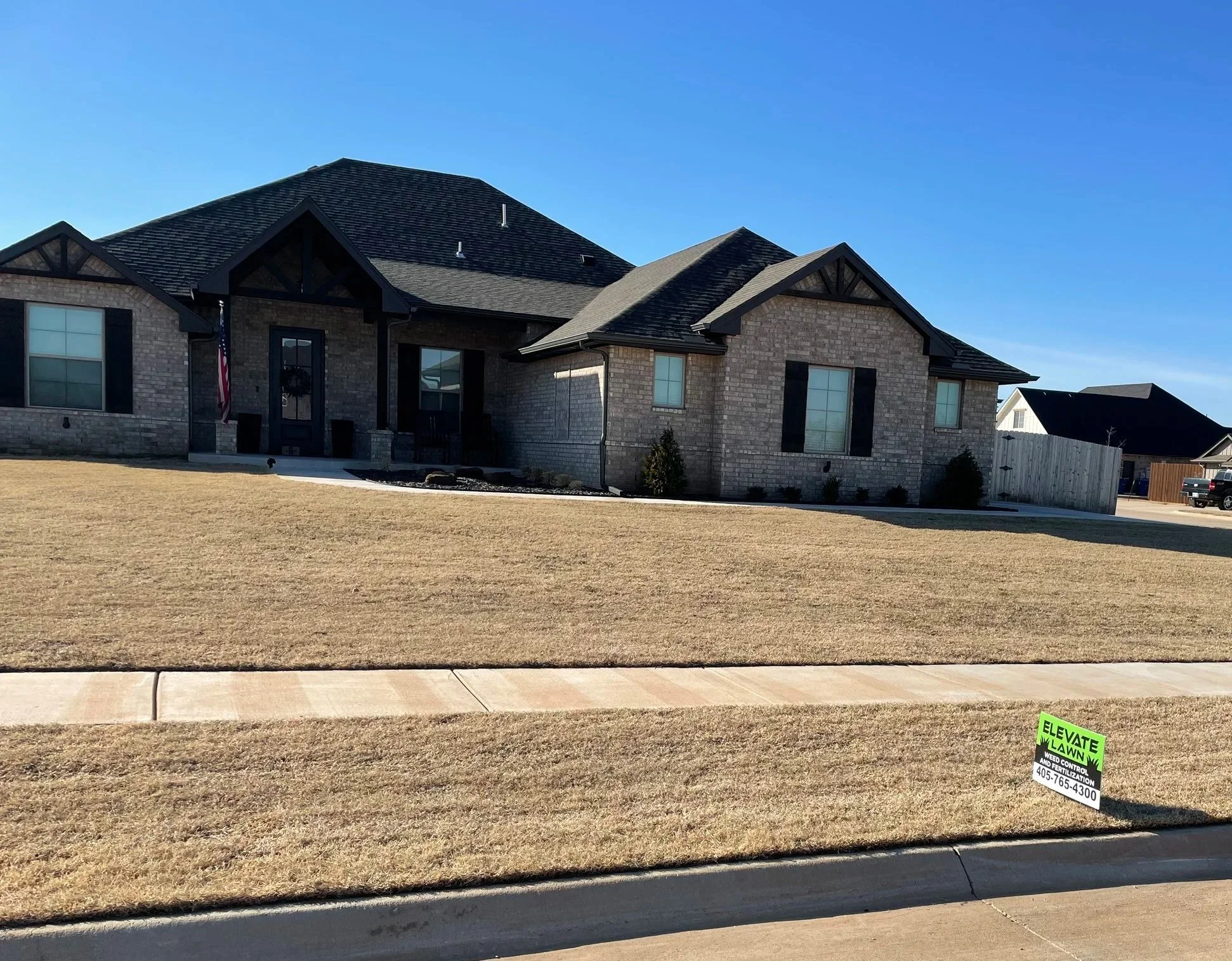 Gray brick suburban house with multiple gabled roofs, front lawn, sidewalk, and Elevate Lawn weed control sign