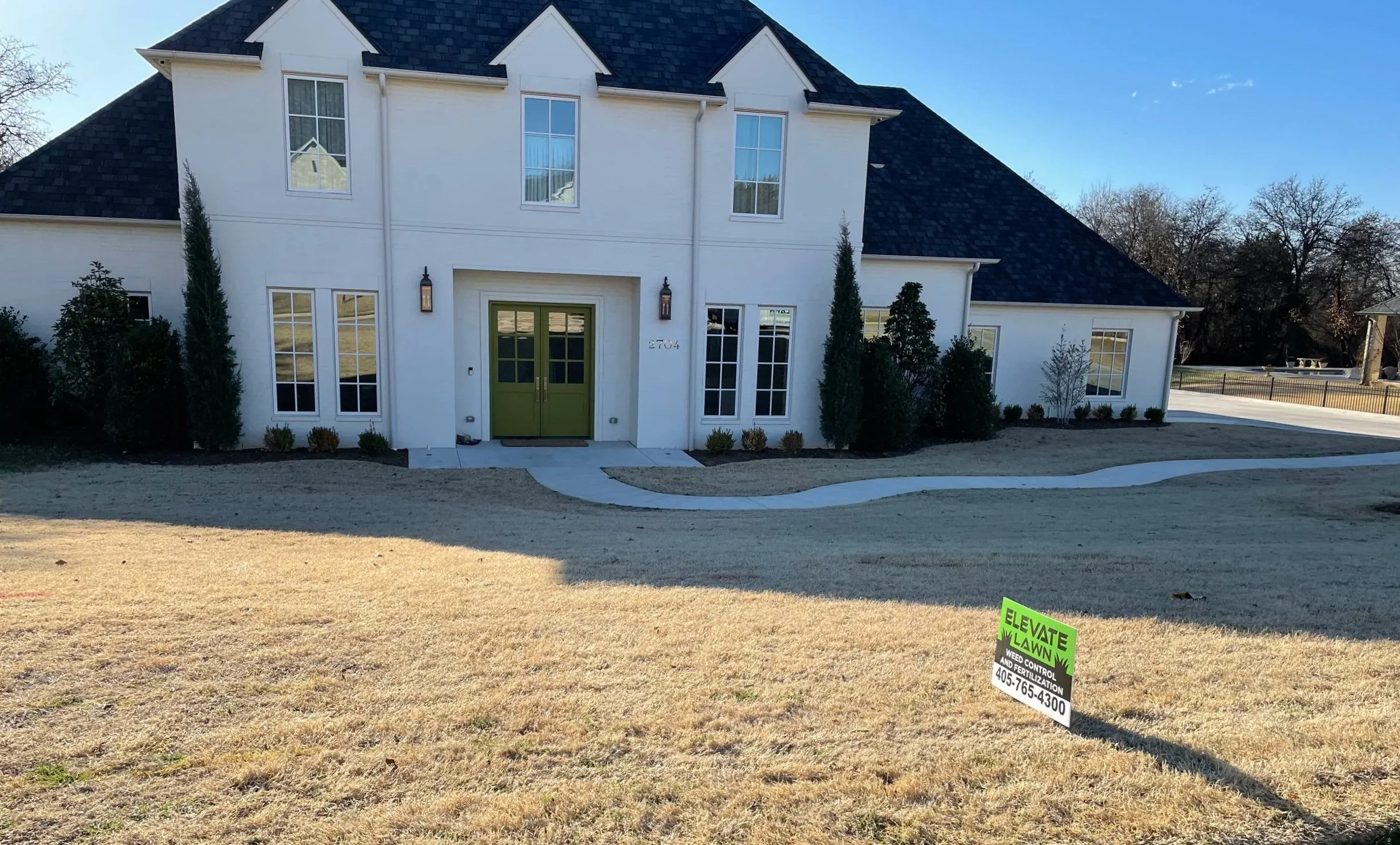 Large white two-story house with dark shingle roof, green double front door, landscaped yard, and lawn treatment sign.