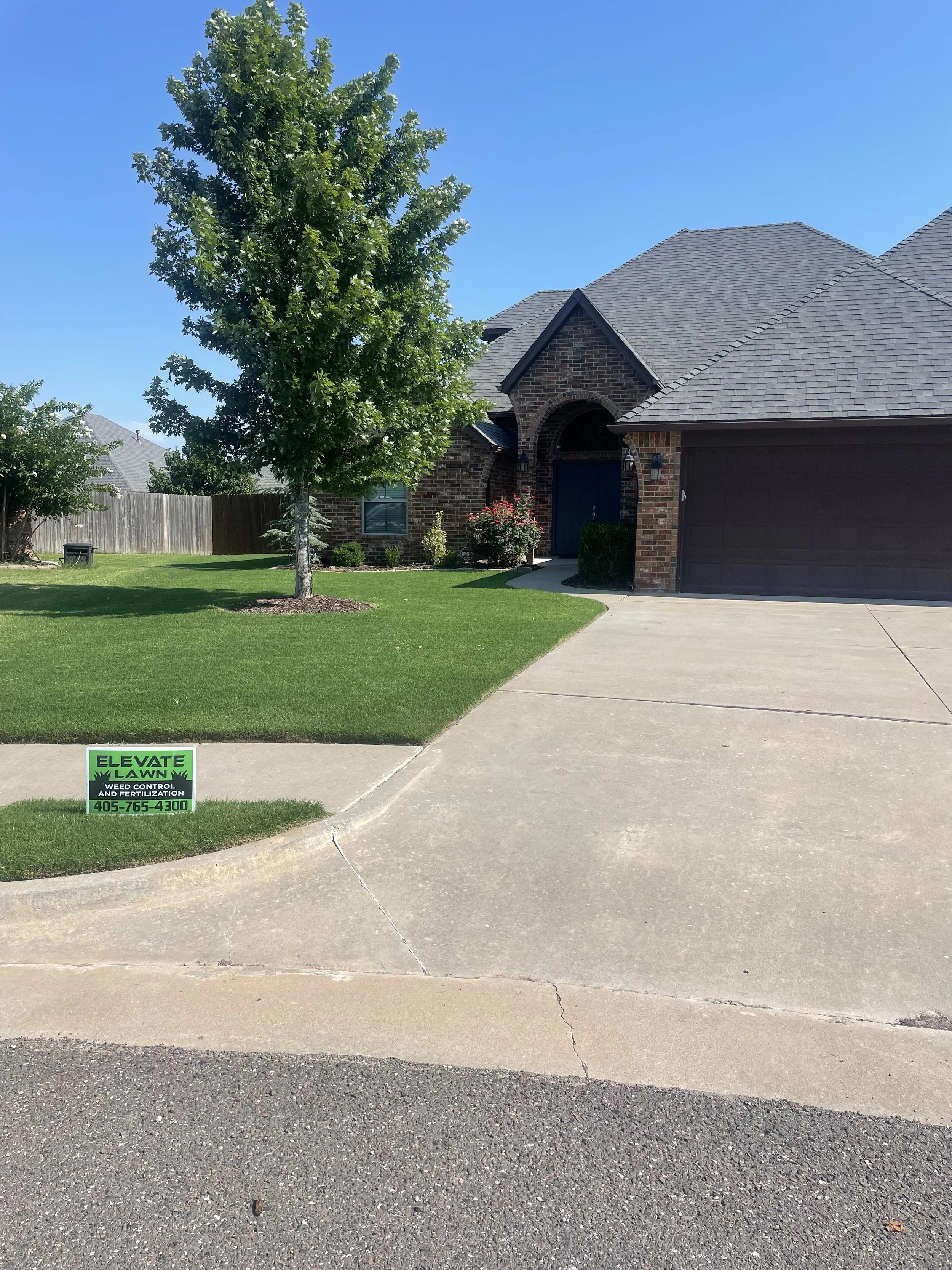 A house with a brick exterior and a gray shingle roof, a well-maintained lawn with a tree, and a driveway leading to a garage. A green yard sign advertises lawn weed control and fertilization services.