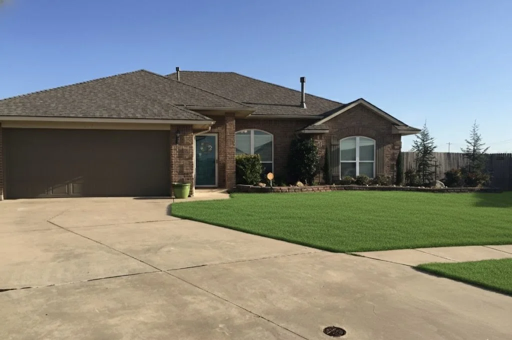 Front yard of a brick house with a well-maintained green lawn, concrete driveway, and a blue front door. A small potted plant is near the entrance, and there are bushes and trees along the house and fence under a clear blue sky.
