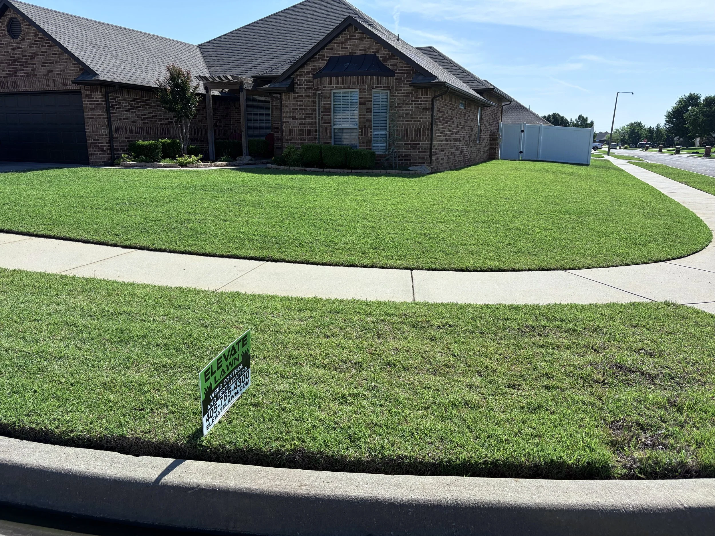 Large brick suburban house with a garage, covered entry, manicured lawn, sidewalk, and lawn-care sign