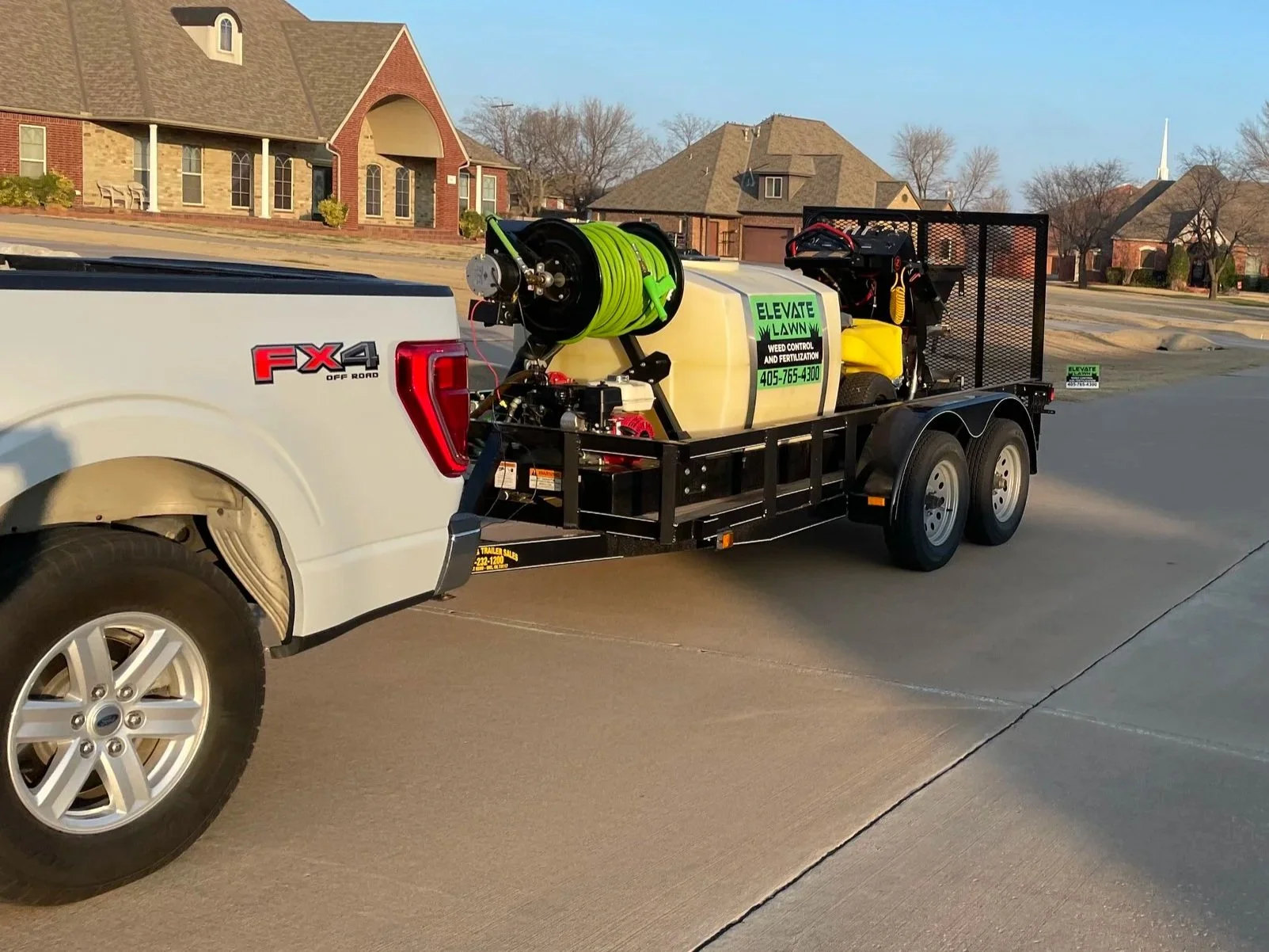 A white Ford pickup truck towing a black utility trailer with a yellow tank, a green hose reel, and landscaping equipment in a residential neighborhood.