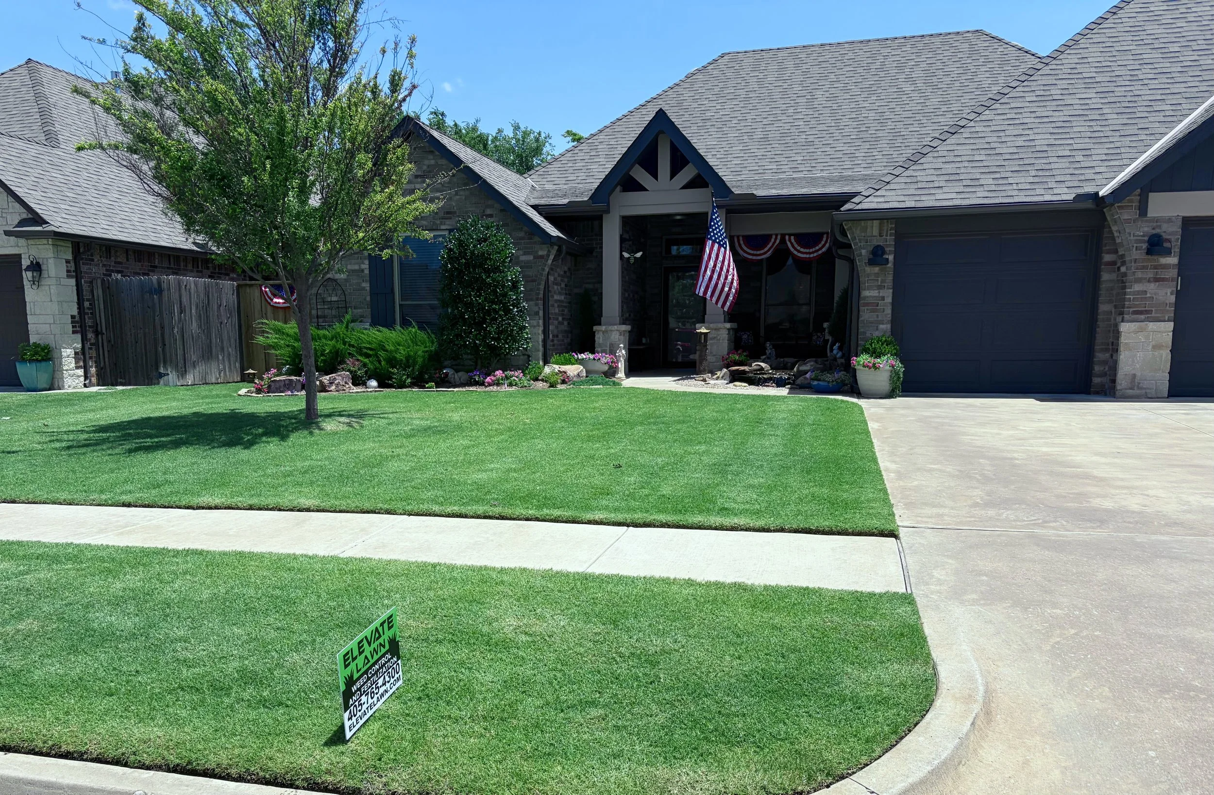 A suburban house with a gray shingled roof and brick exterior, front lawn with green grass and flowers, American flag hanging by the door, and a sidewalk in the foreground with a green sign that reads 'ELEVATE LAWN'.