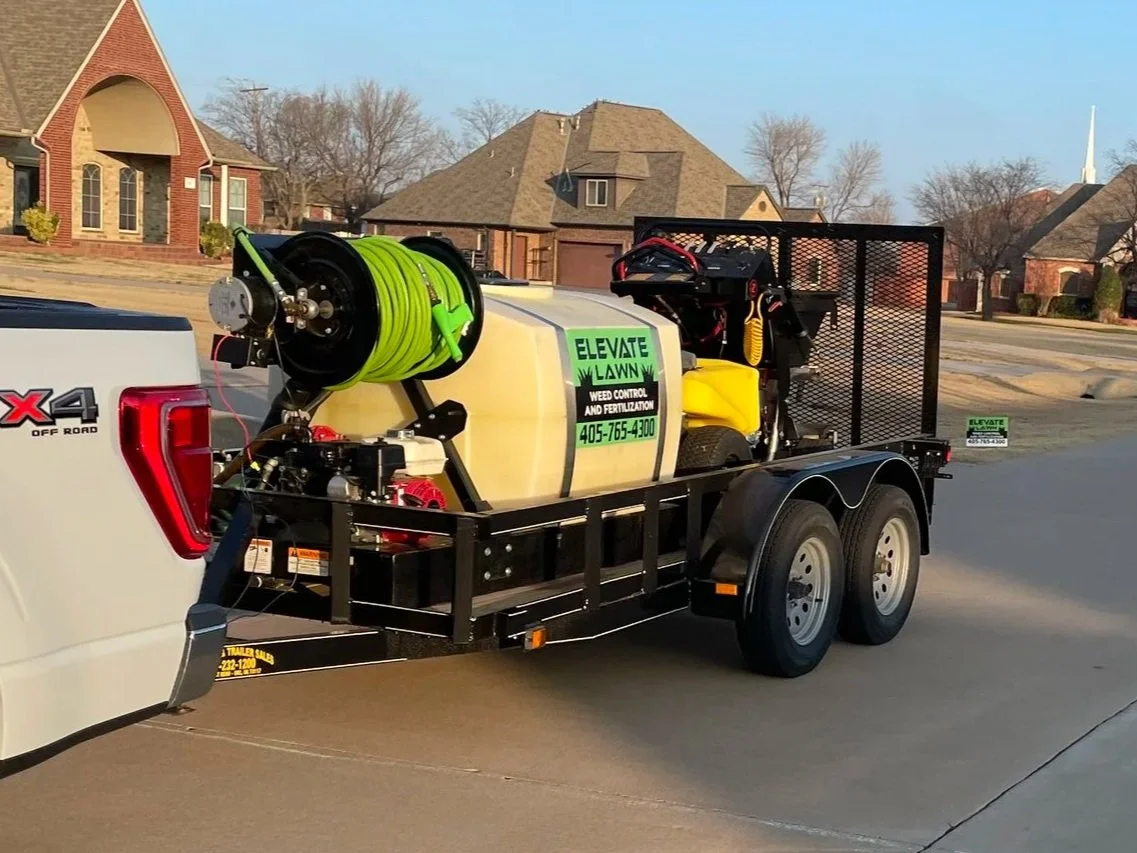 Trailer attached to a pickup truck labeled 'Elevate Lawn Weed Control and Fertilization,' with equipment including a large green hose reel, motorized pump, and other yard maintenance gear, parked on a suburban street with houses in the background.