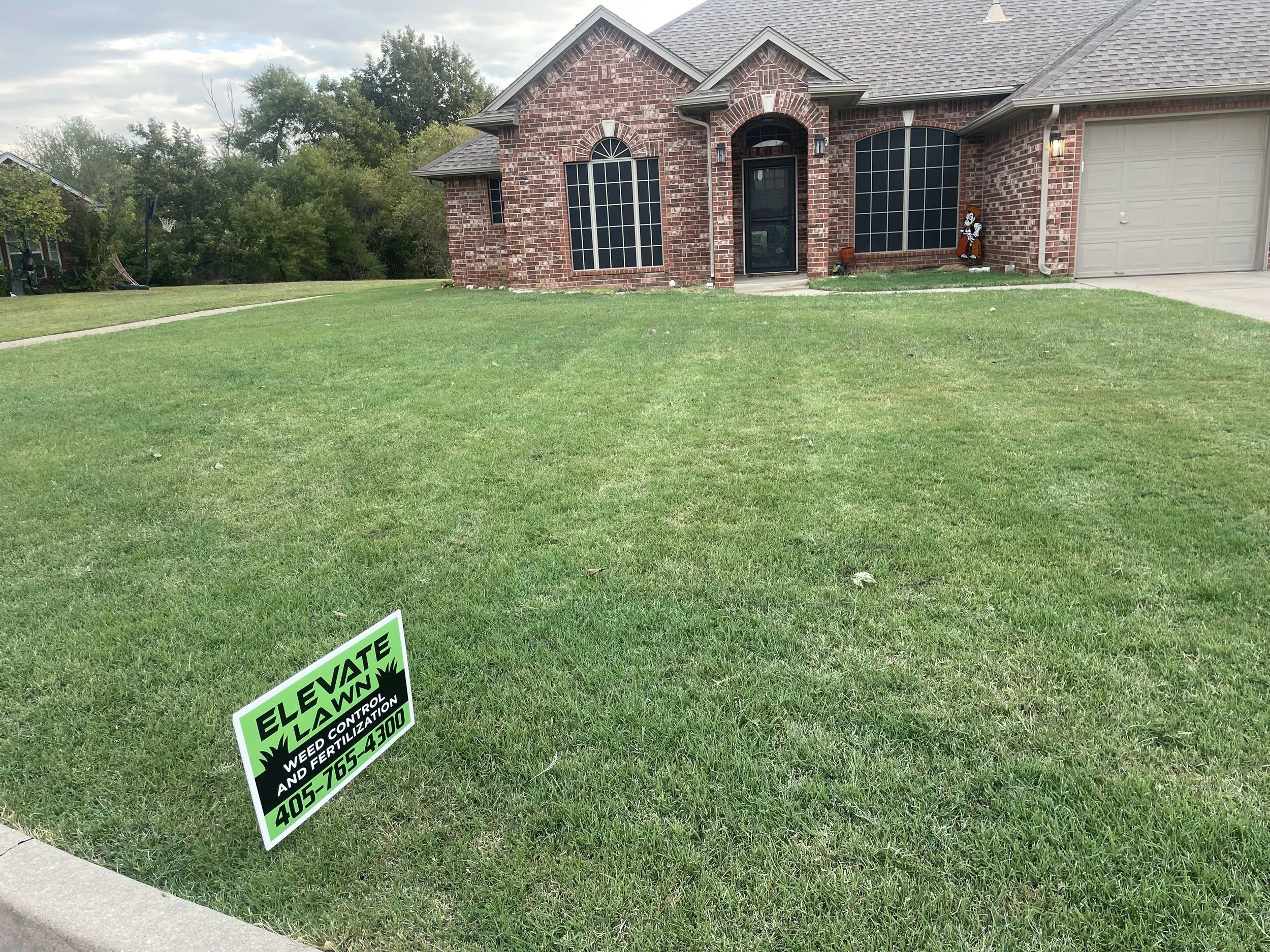 Front yard of a brick house with a green lawn, a sign in the grass that reads 'ELEVATE LAWNS, WEED CONTROL AND FERTILIZATION, 405-765-4300', a driveway, and a small decorative figure near the house.