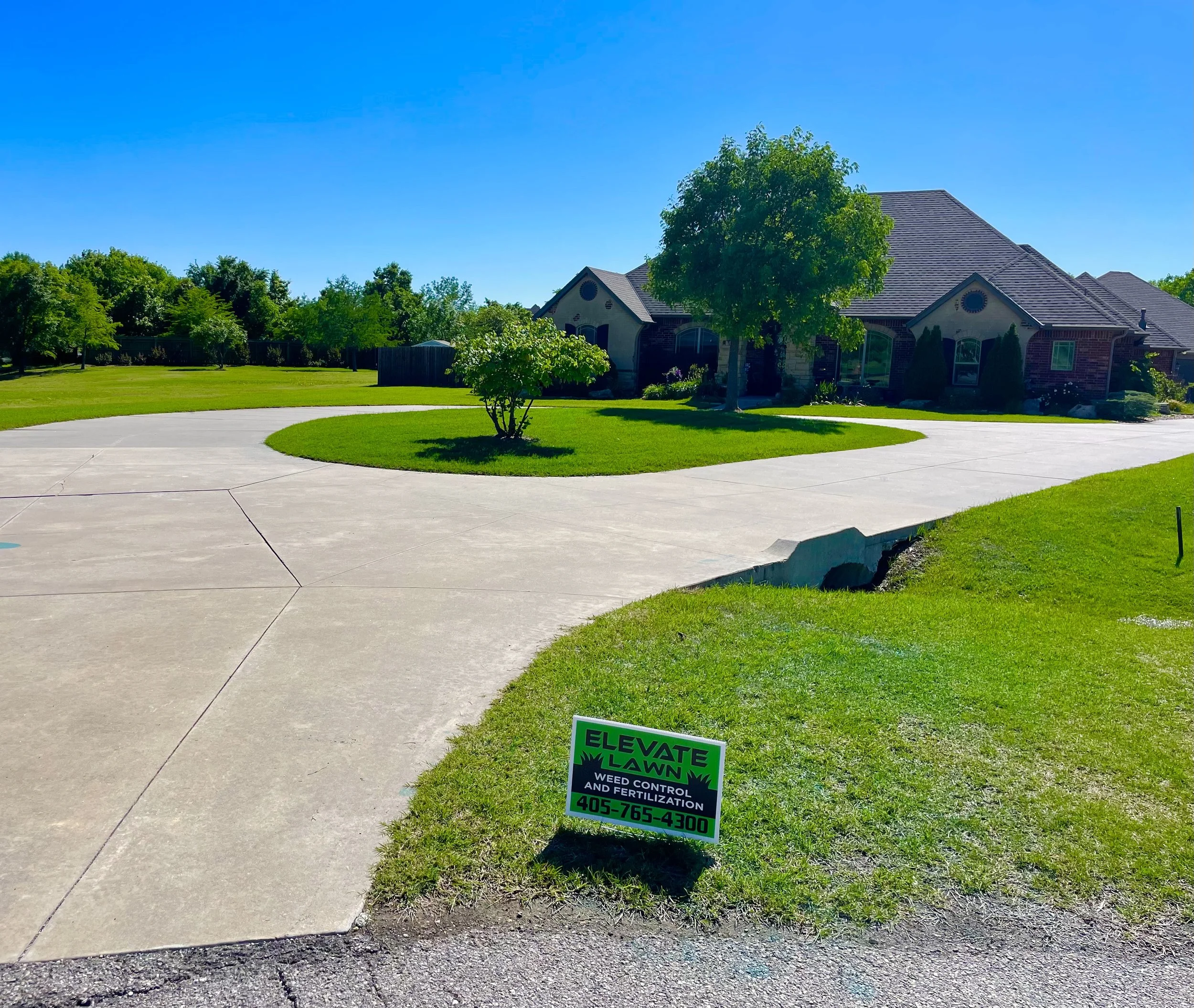 A large brick house with a dark roof, surrounded by green trees and lawn, along with a curved concrete driveway. A small sign in the front yard reads Lose K-9 weed control and fertilization, with a phone number.