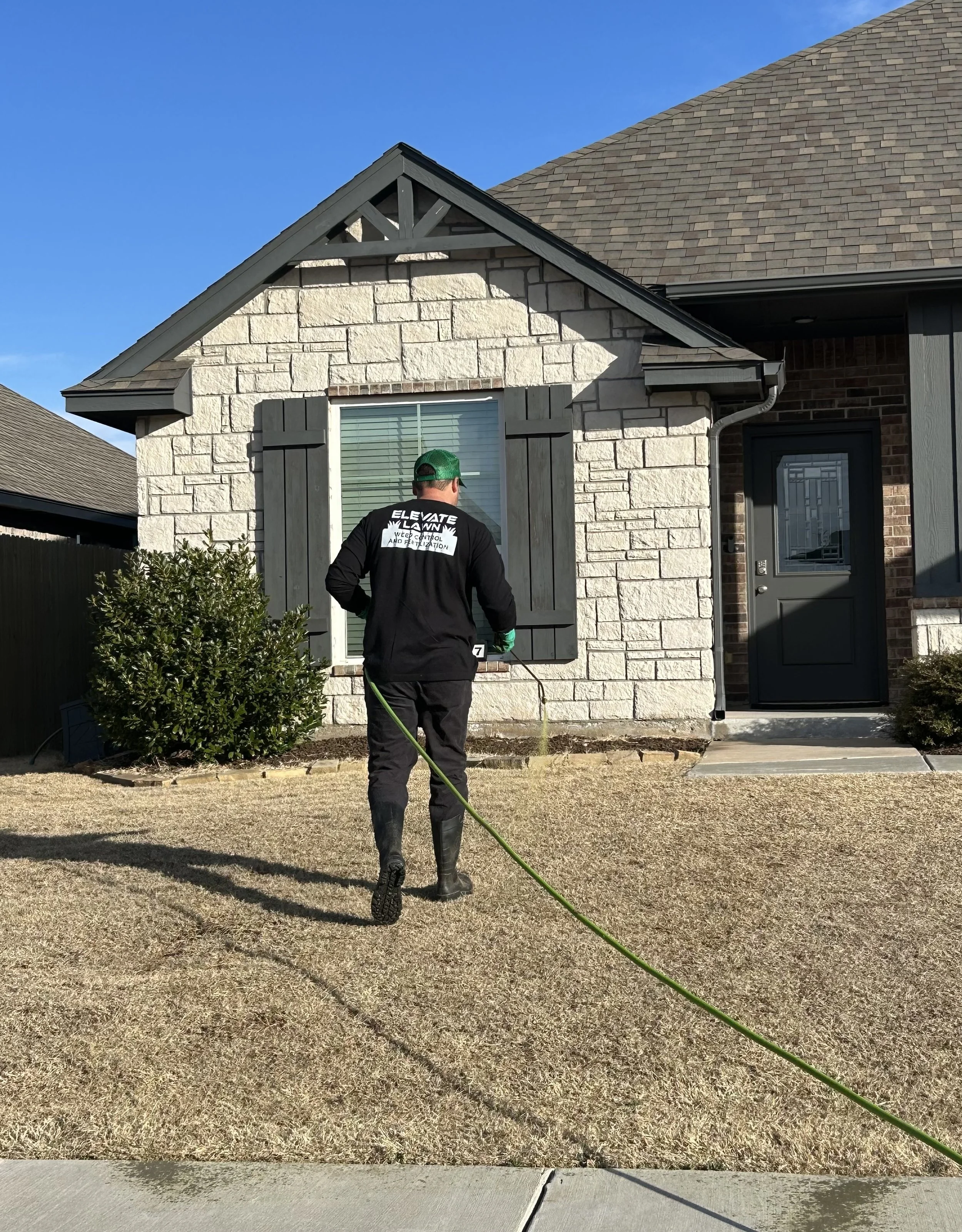 A person in work clothes and boots spraying pre-emergent on a front yard of a house with a hose. The house has a stone facade and dark shutters, with a tree and bushes in the yard. The sky is clear and blue.