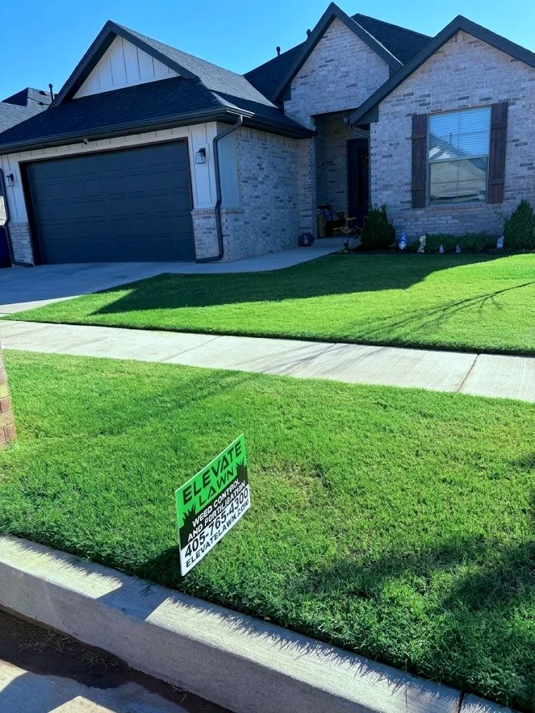 Front yard of a house with well-manicured green lawn, concrete walkway, and a house with brick exterior and dark-colored roof. There is a green sign on the grass advertising lawn weed control and insect control services with a phone number and website.