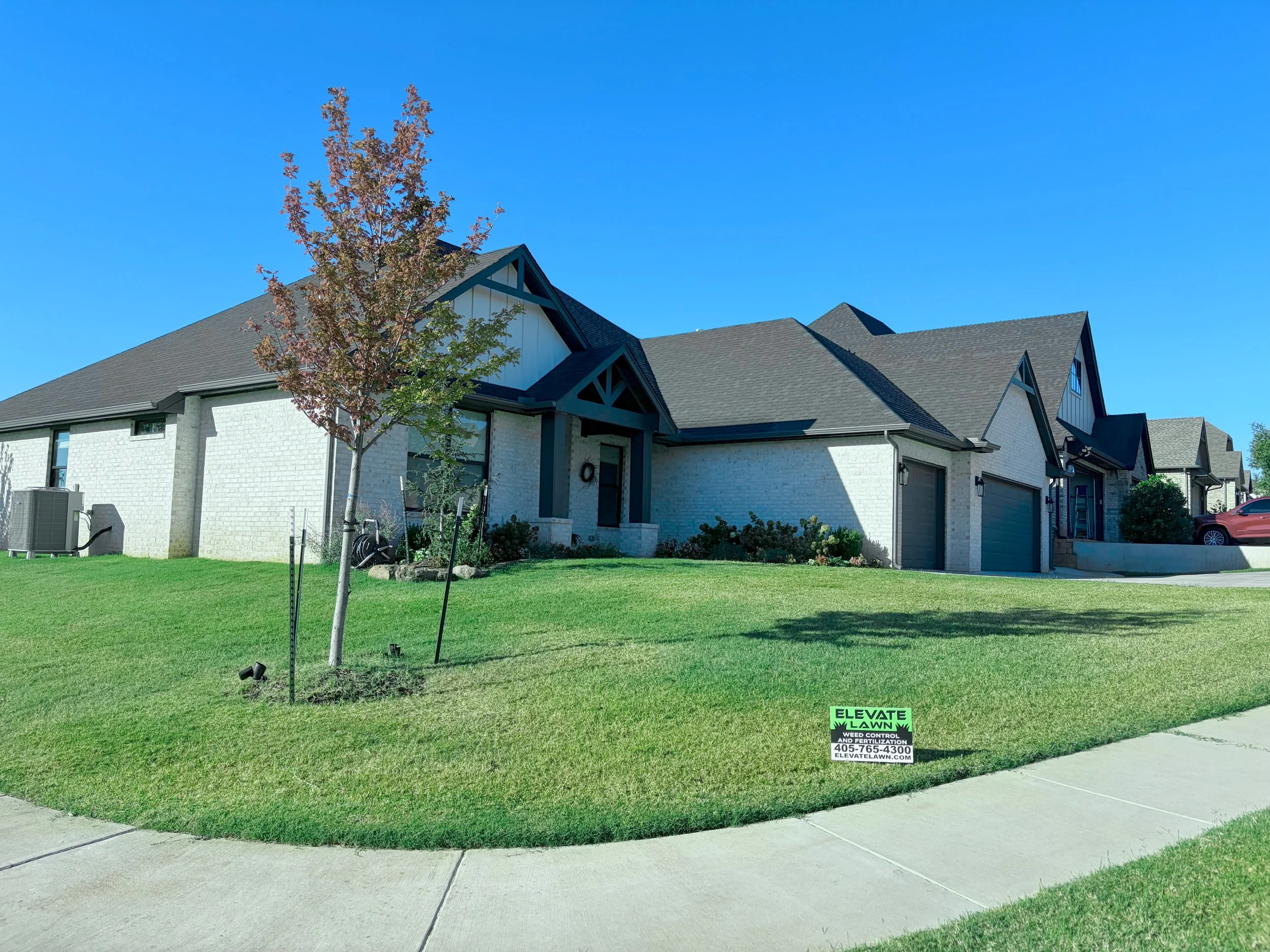 Well-maintained suburban home with white brick exterior, manicured lawn, young tree, and Elevate Lawn weed control sign.