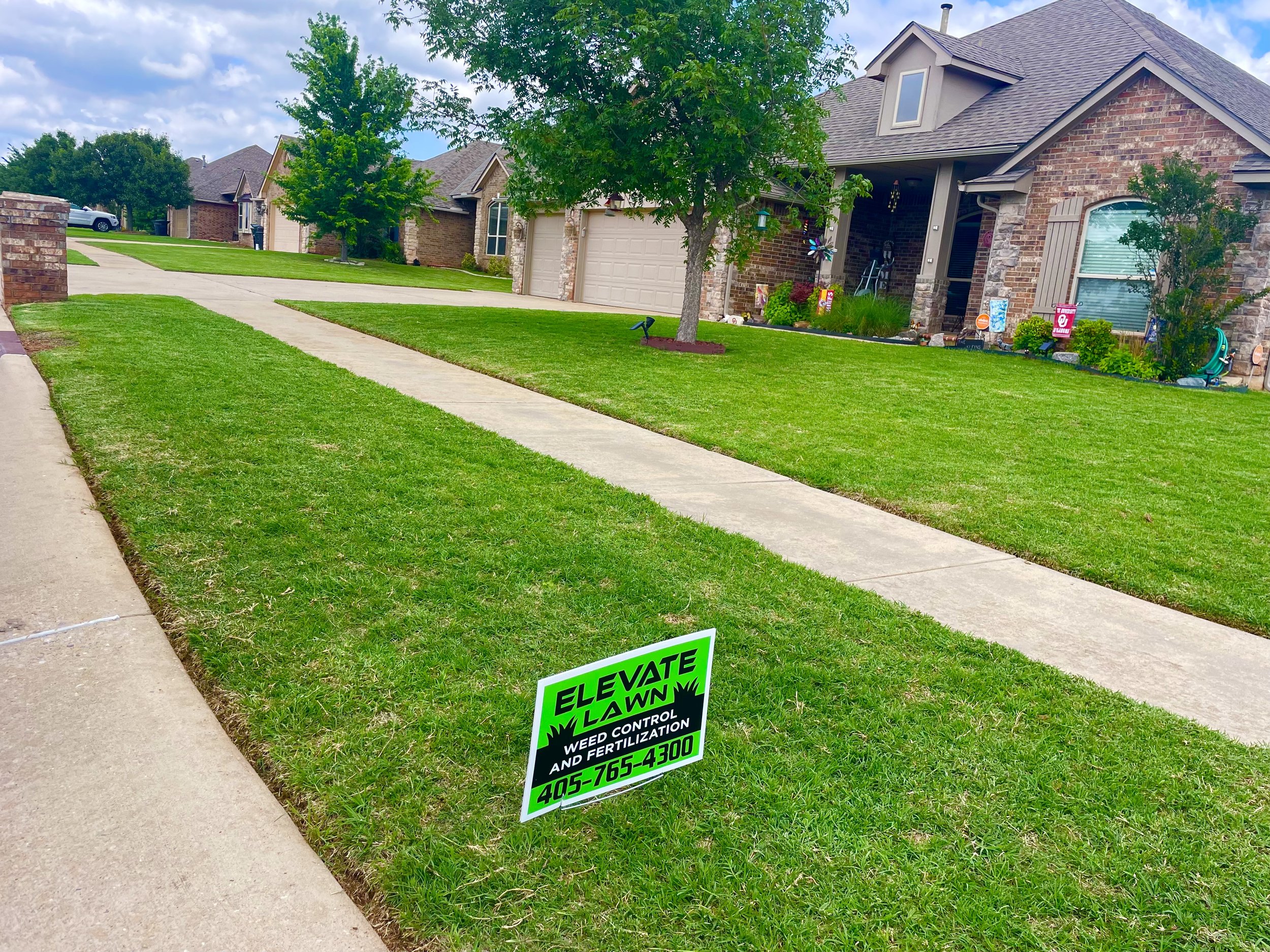 Front yard with a sidewalk, green grass, a tree, and a house with a brick exterior. A yard sign for "Elevate Lawn" indicates weed control and fertilization services, including a contact phone number.