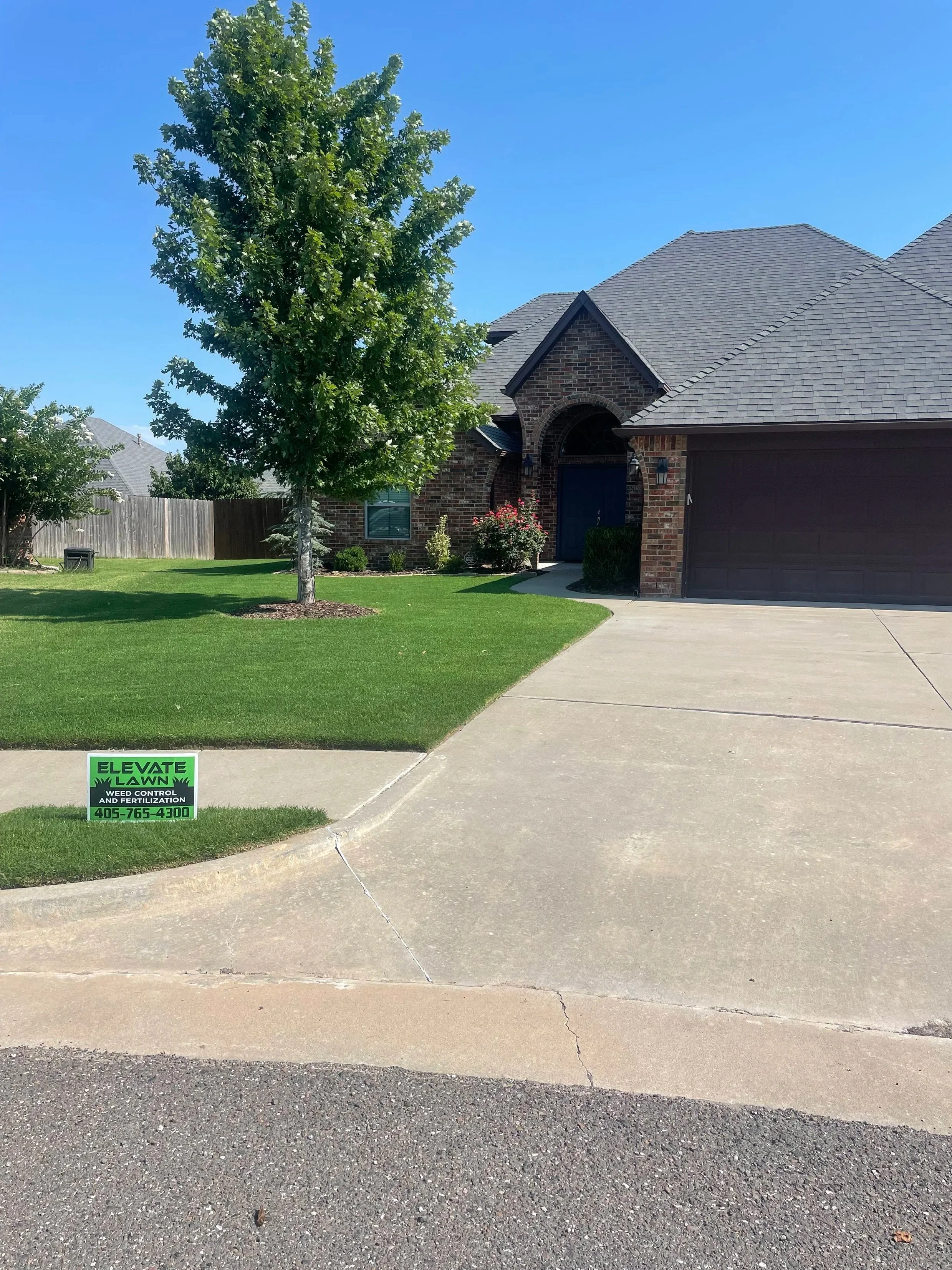 A suburban house with brick exterior, a large tree in the front yard, a well-maintained lawn, and a concrete driveway leading to a two-car garage. A small green sign advertising weed control and fertilization is in the grass.